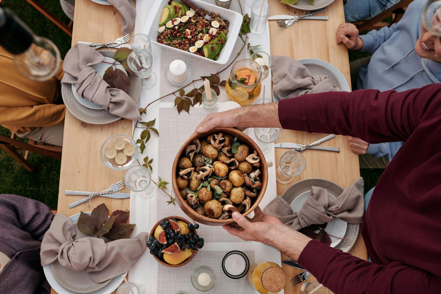 Overhead view of a dinner party table with shared plates of food, wine glasses, and hands reaching for dishes.