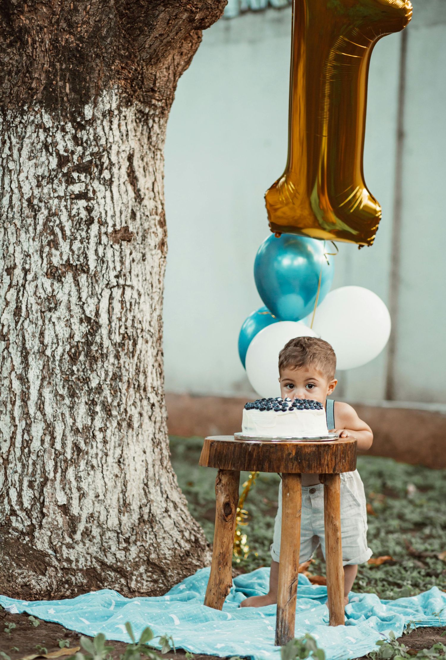 A first birthday celebration setup with gold number one balloon, blue balloons, and a rustic wooden stool on blue fabric. Anthem Arizona.