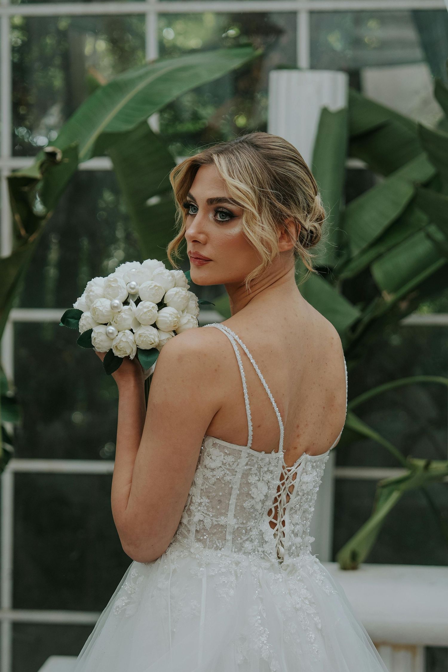 A bride in a white lace dress poses with white roses against a greenhouse backdrop. Leading Arizona Photographer.