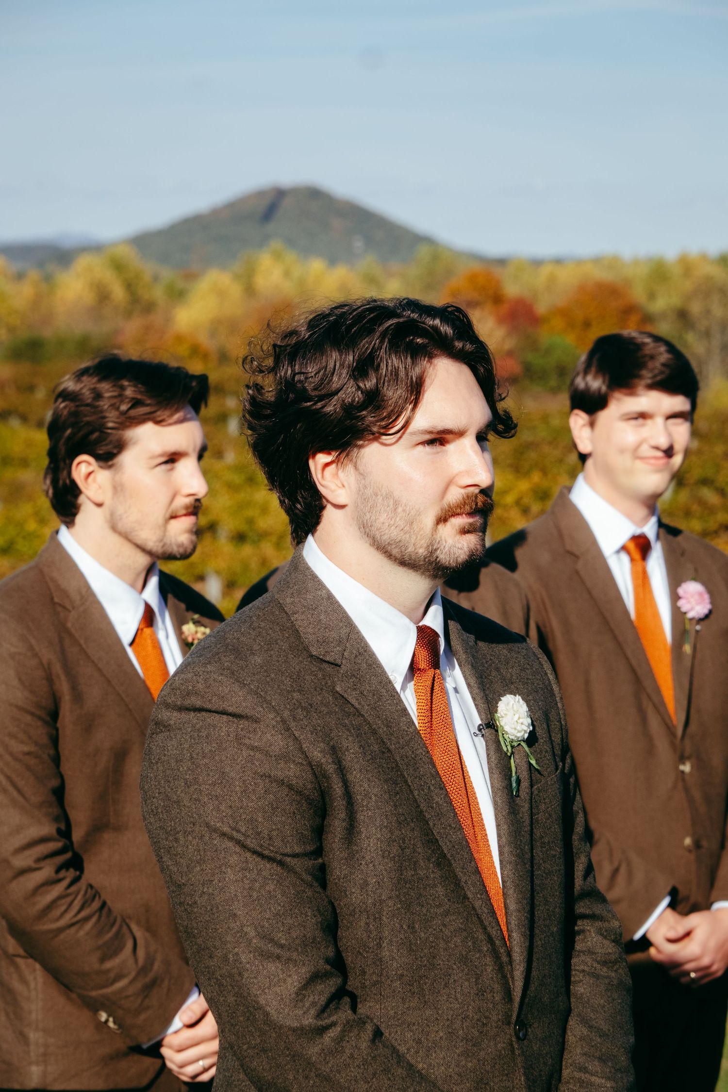Groomsmen wearing brown suits with orange ties stand in a line against an autumn landscape background.