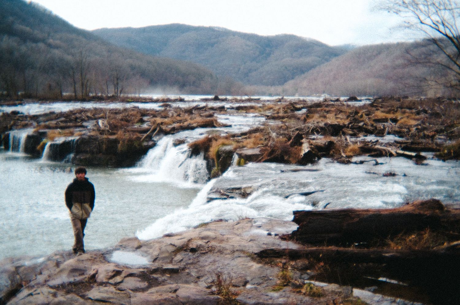 Person standing near frozen waterfall cascading over rocky terrain in winter mountain landscape.