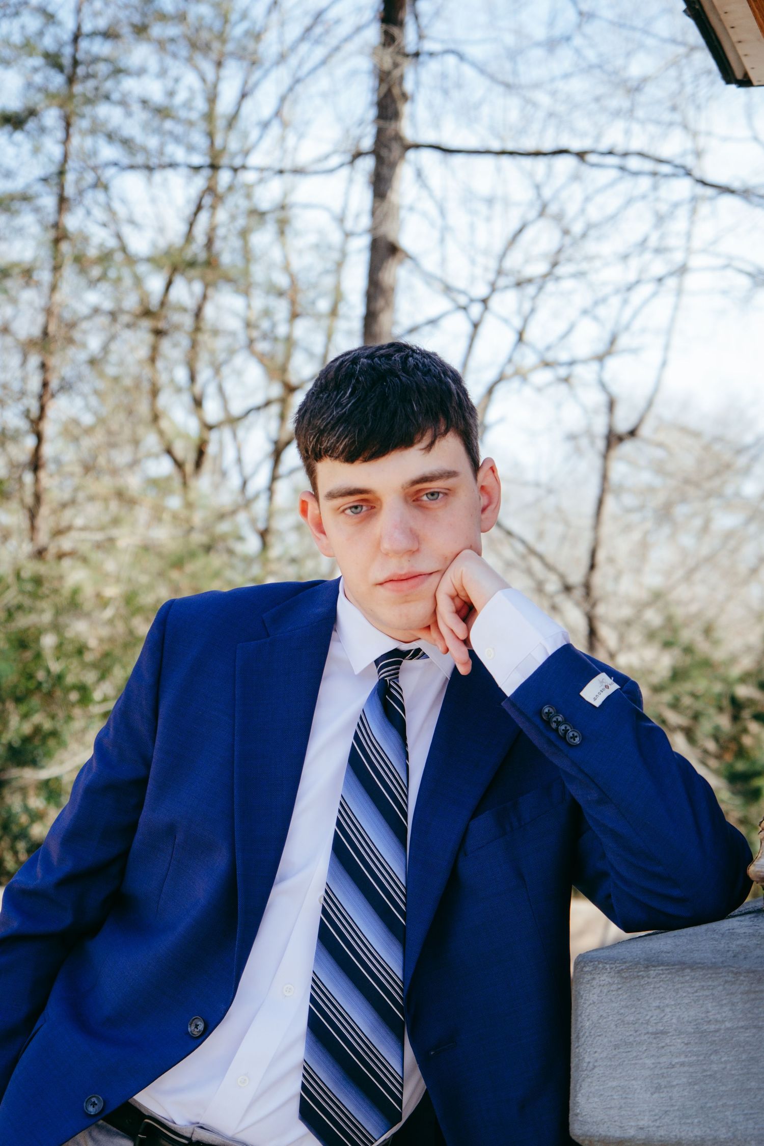 A person in a navy suit and striped tie rests their head on their hand while sitting outdoors.