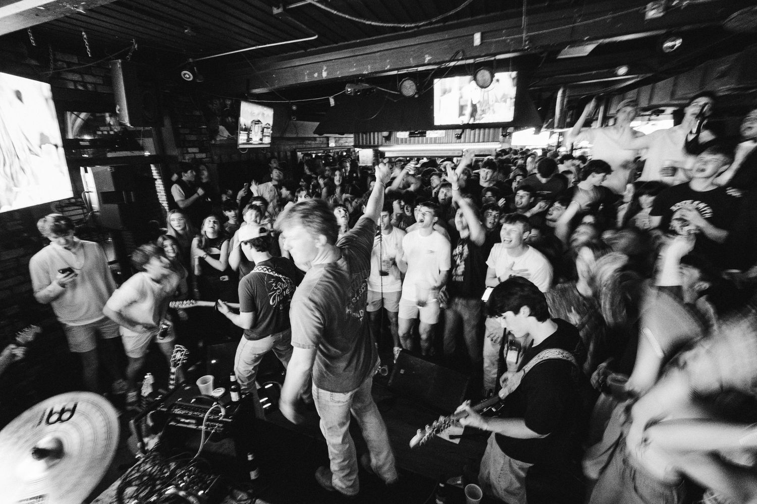 Energetic crowd dancing and cheering at a packed indoor concert venue with dark lighting and stage setup.
