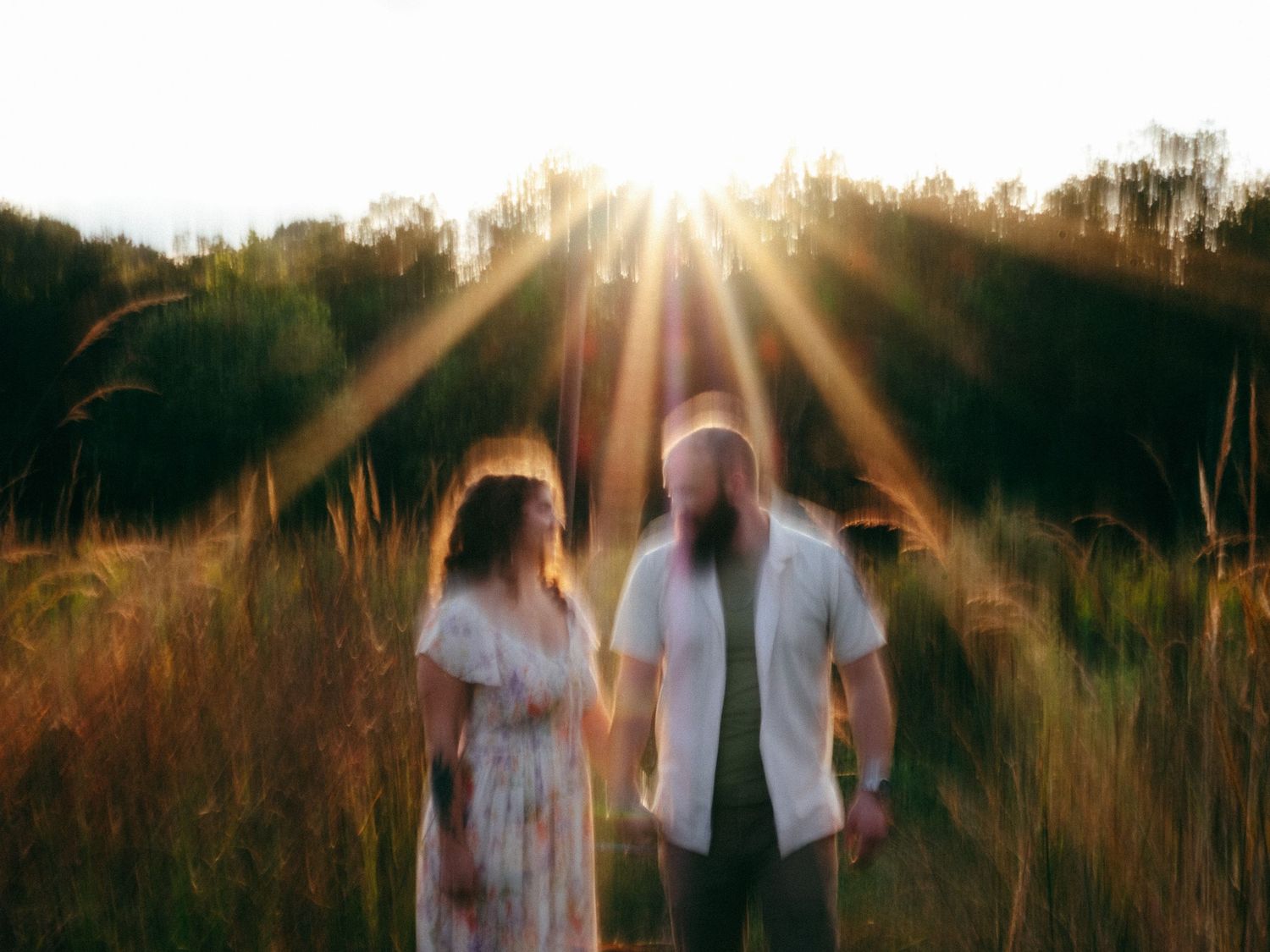Couple walking through golden sunlit field at sunset with lens flare and tall grass creating a romantic atmosphere.