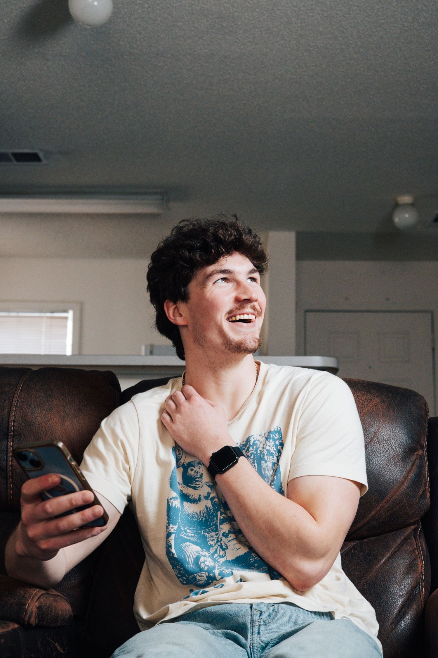 A person in denim overalls and white t-shirt sits on a brown leather couch while looking at their phone and laughing.