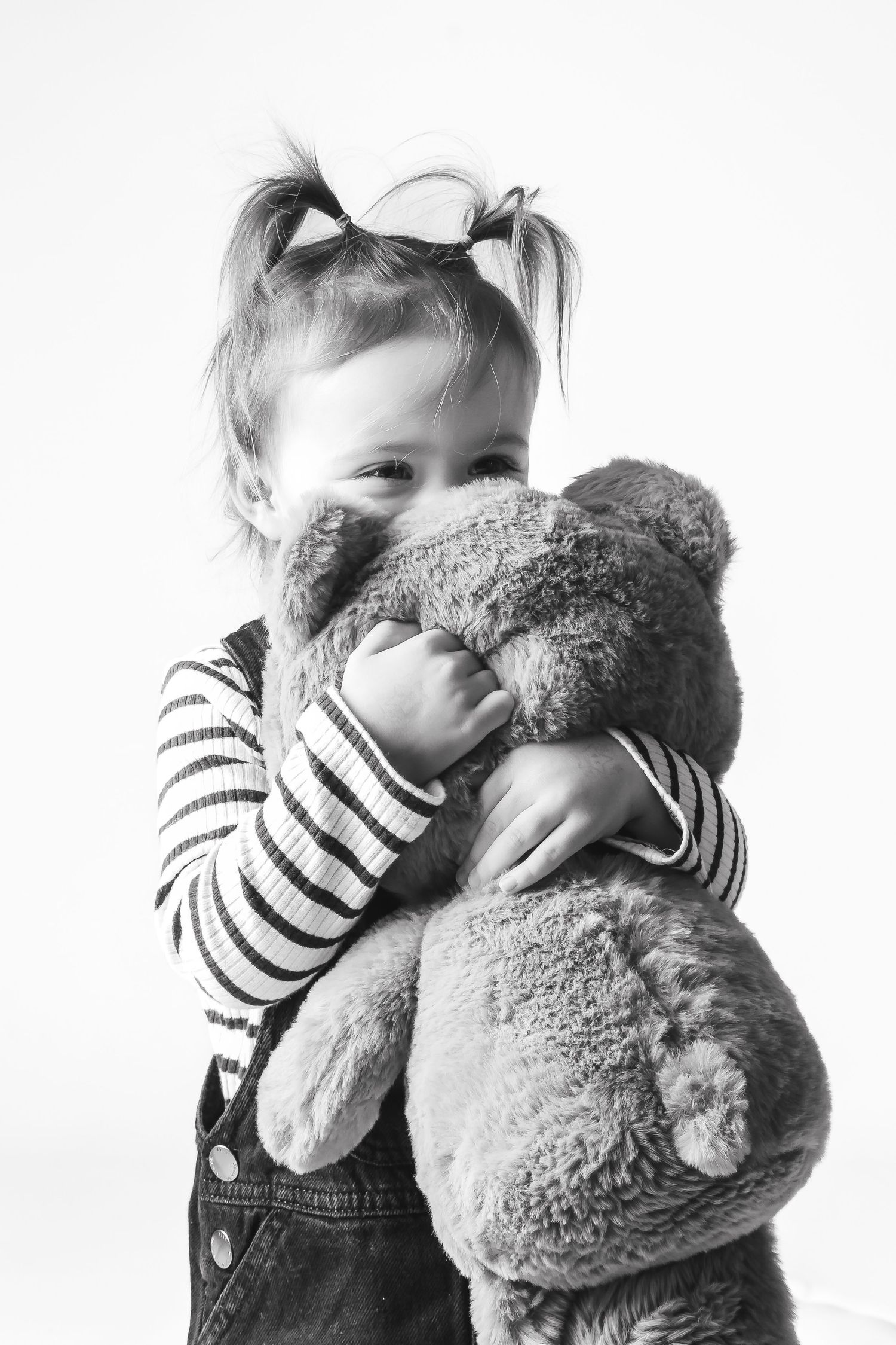 Black and white photo of a small child in striped shirt and overalls hugging a plush teddy bear.