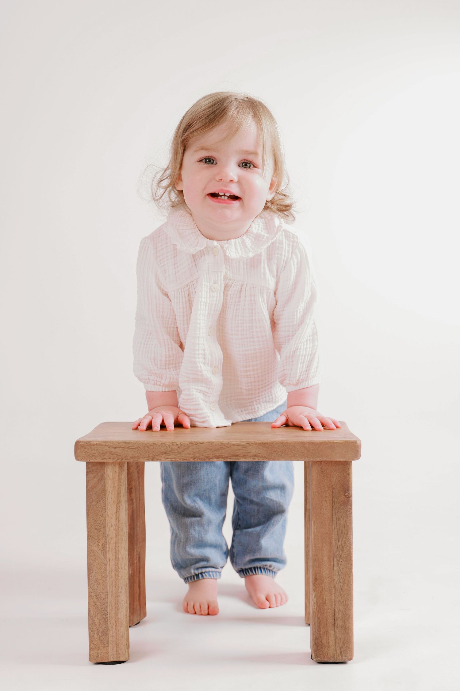 Baby in white shirt and jeans sitting on wooden bench showing playful expression.