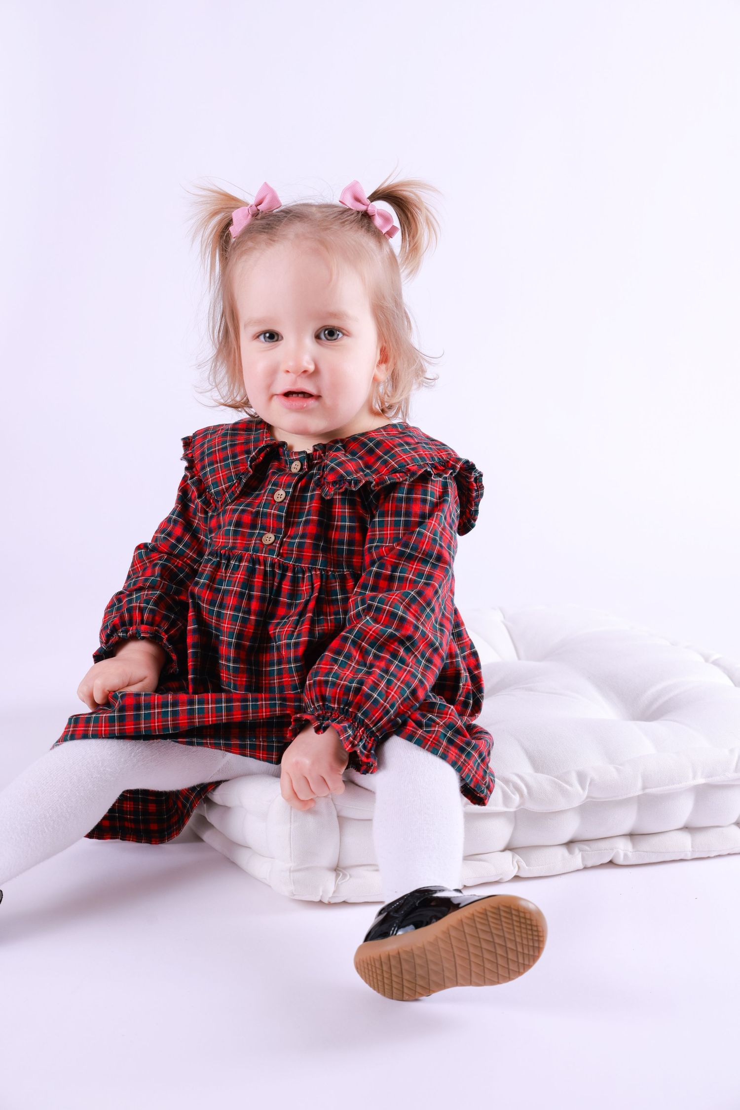 A small child wearing a red plaid dress sits on white bedding while wearing casual shoes.