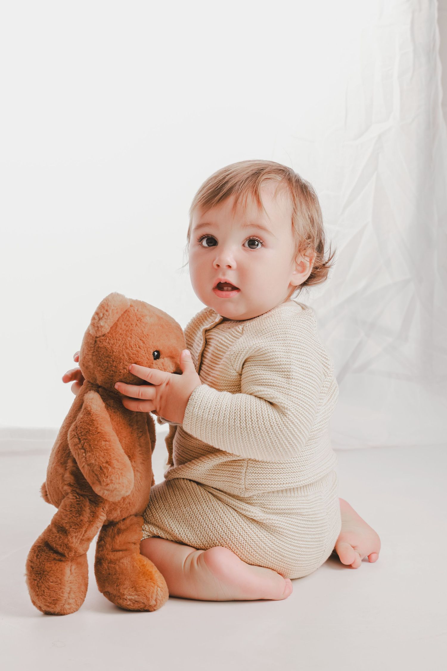 Baby in cream sweater plays with brown teddy bear on white background.