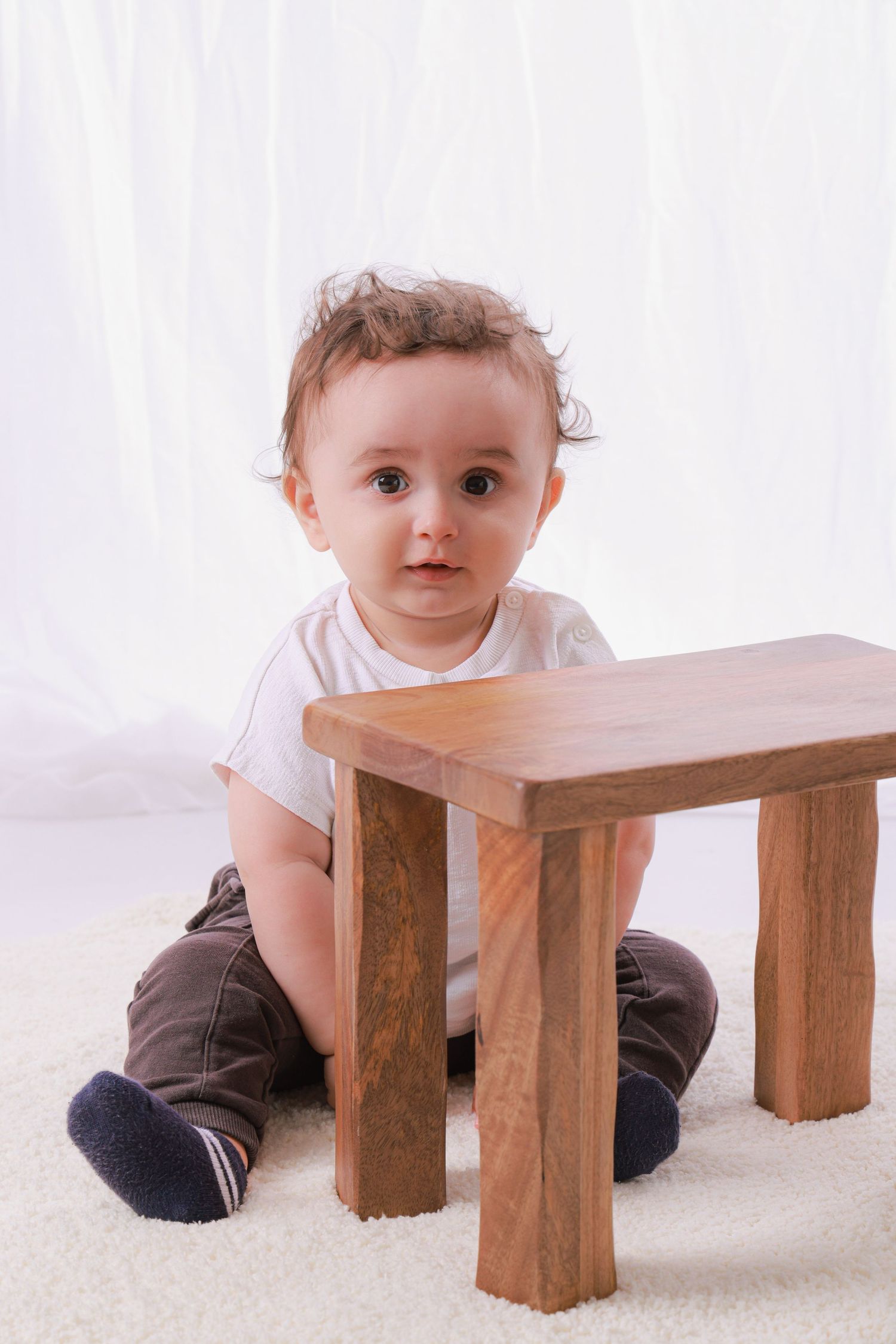 Baby in white shirt crawls around wooden stool on white carpet.
