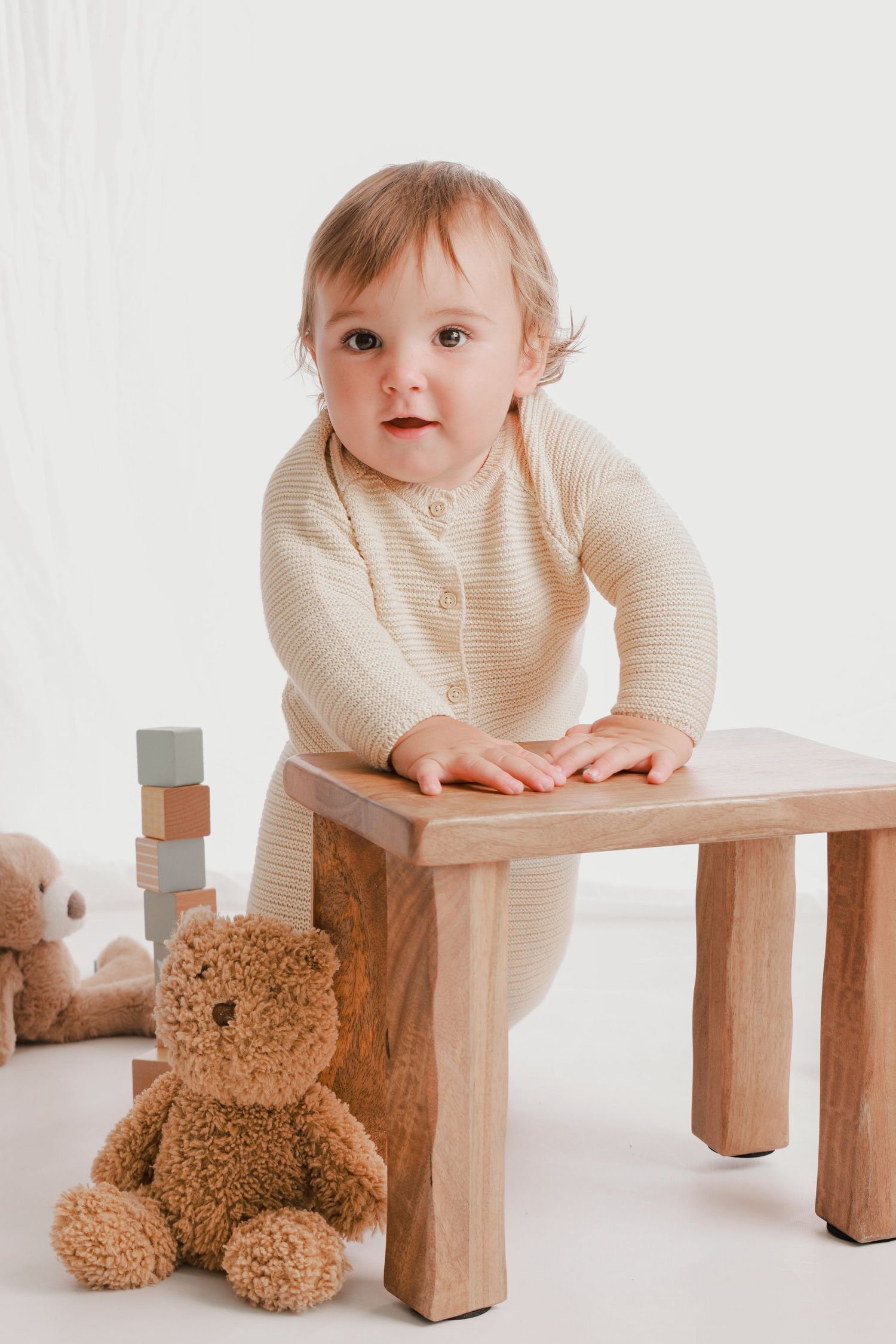 Baby playing with teddy bear and wooden blocks on small bench in studio setting.