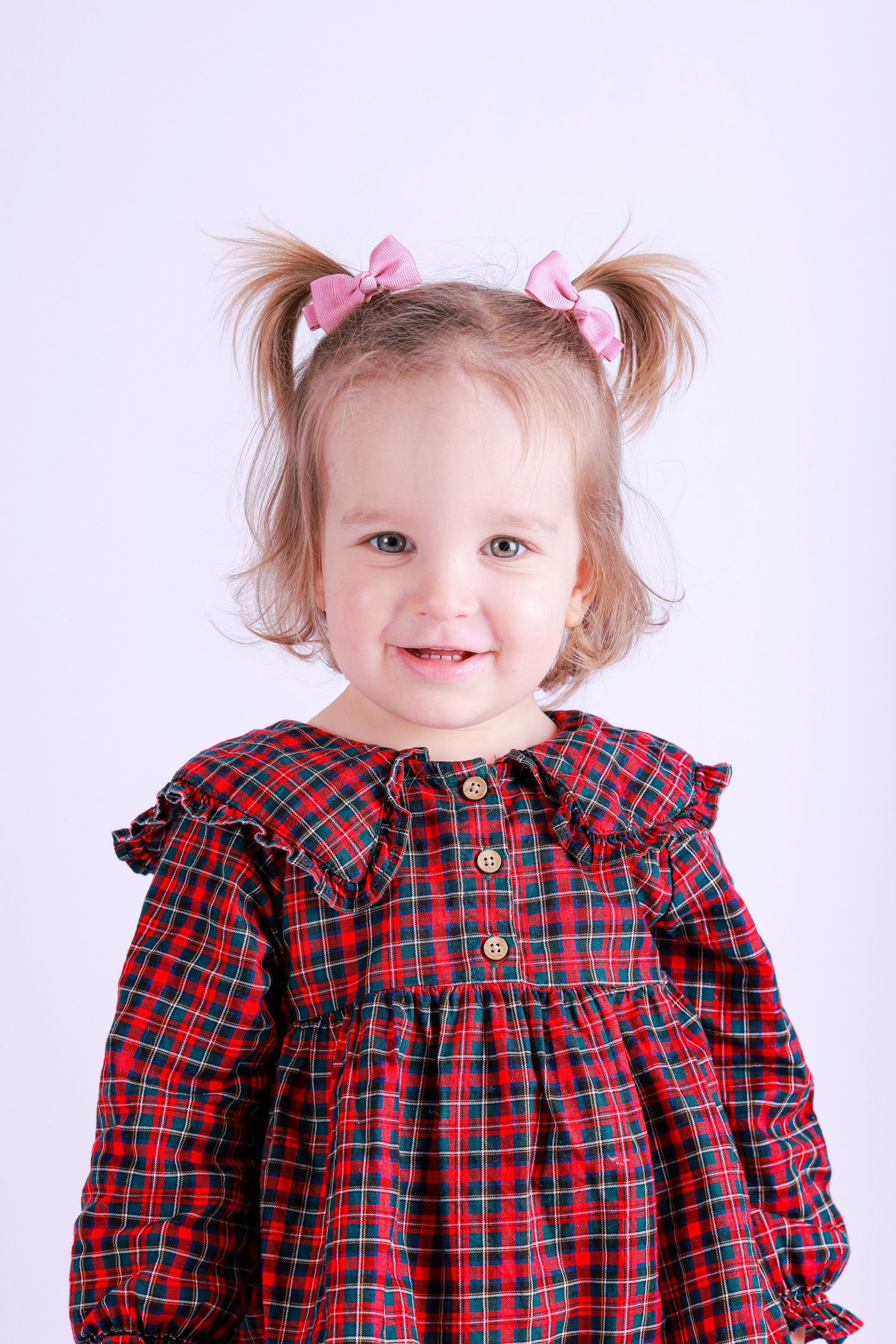A young child in a red plaid dress with pink hair bows smiles while posing for three sequential photos.