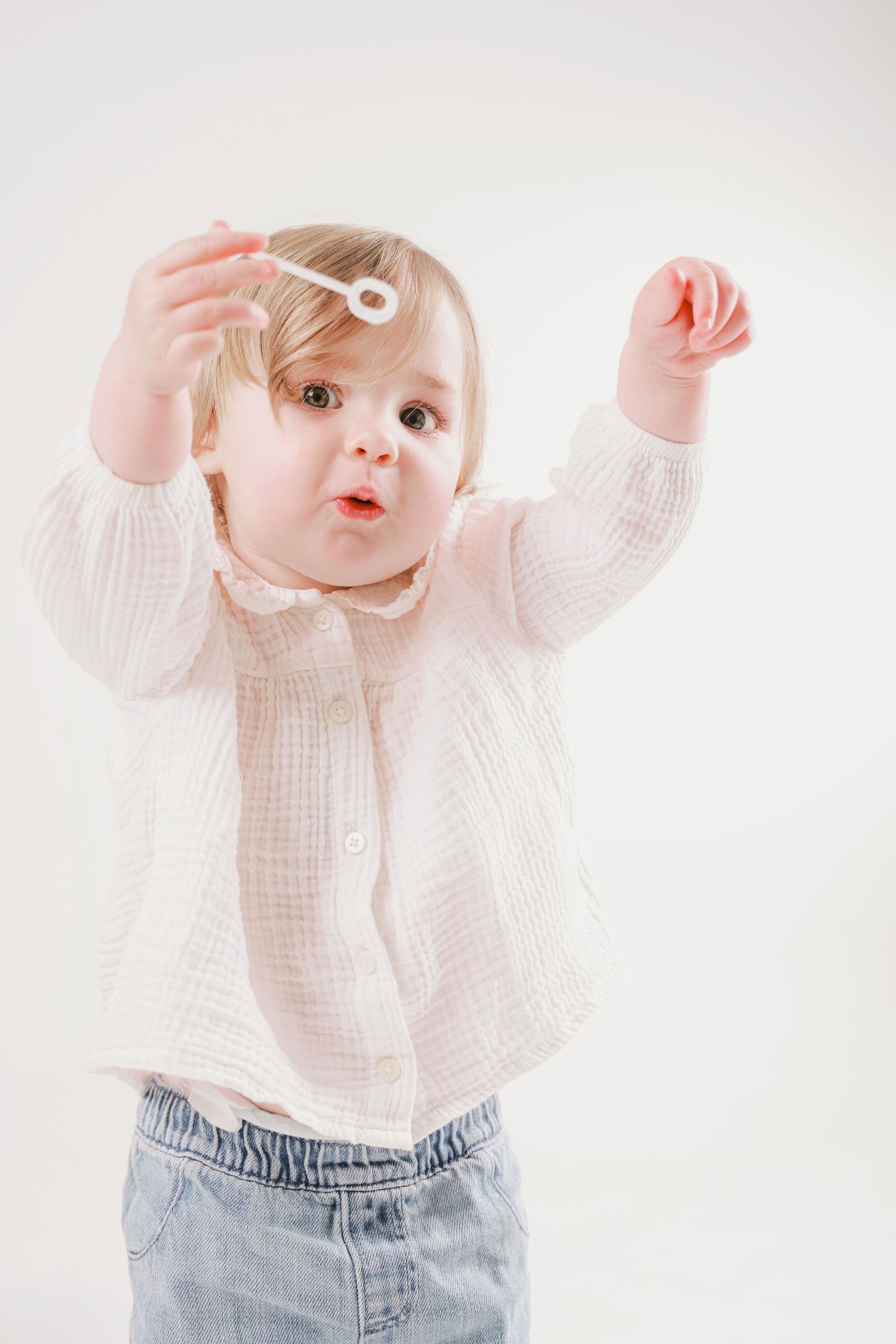 Baby wearing white top and jeans reaching upwards against white background.