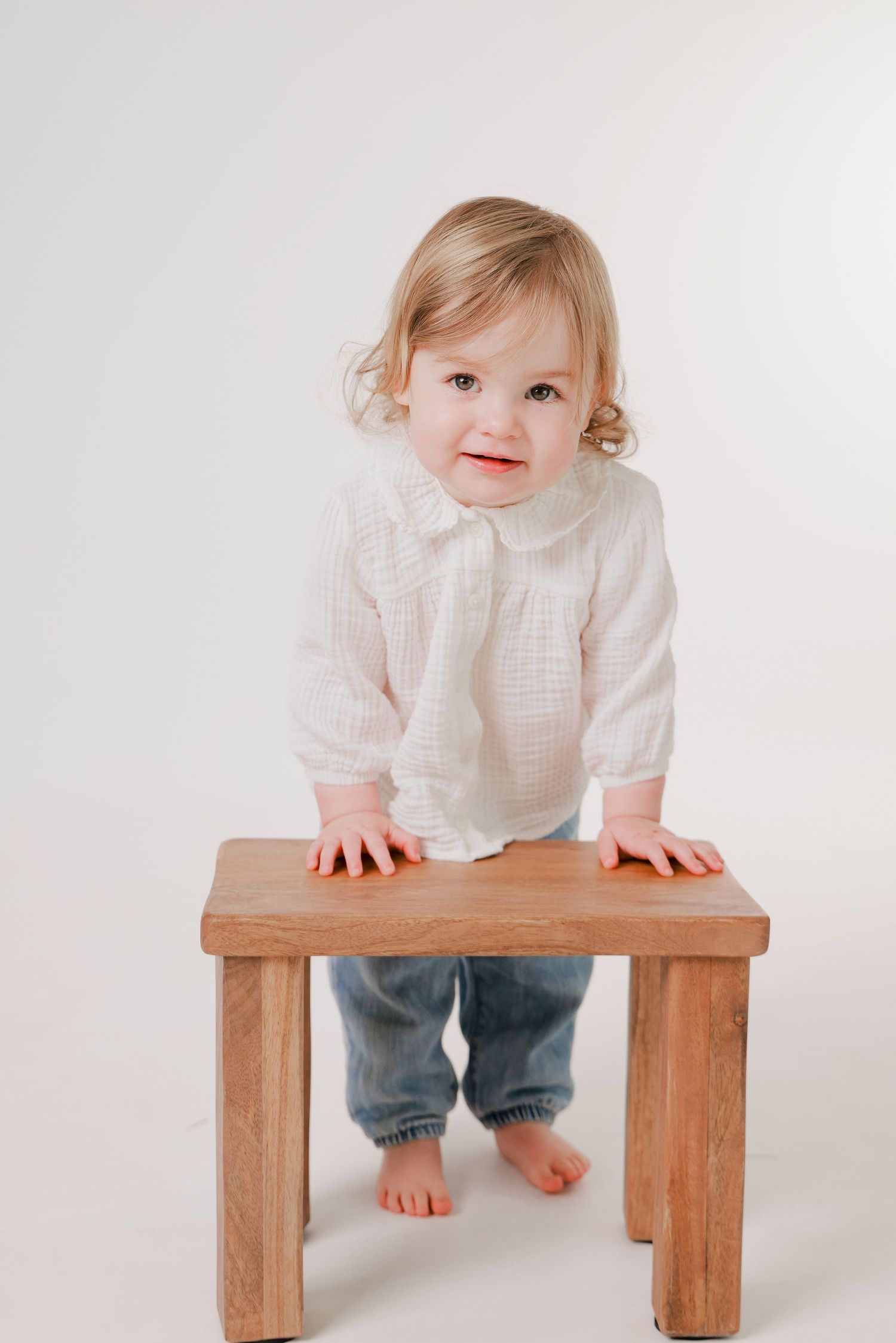 Baby in white shirt explores wooden bench against white background.