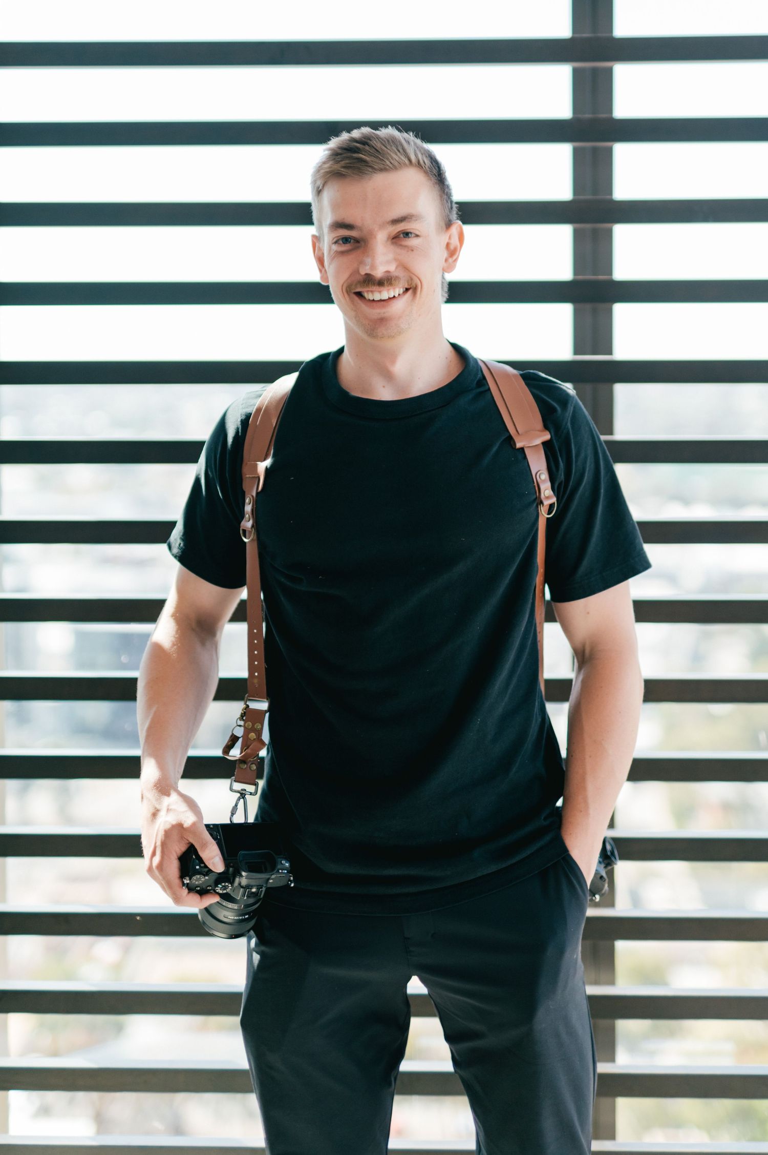 Professional photographer wearing black t-shirt and leather camera straps against striped wall.