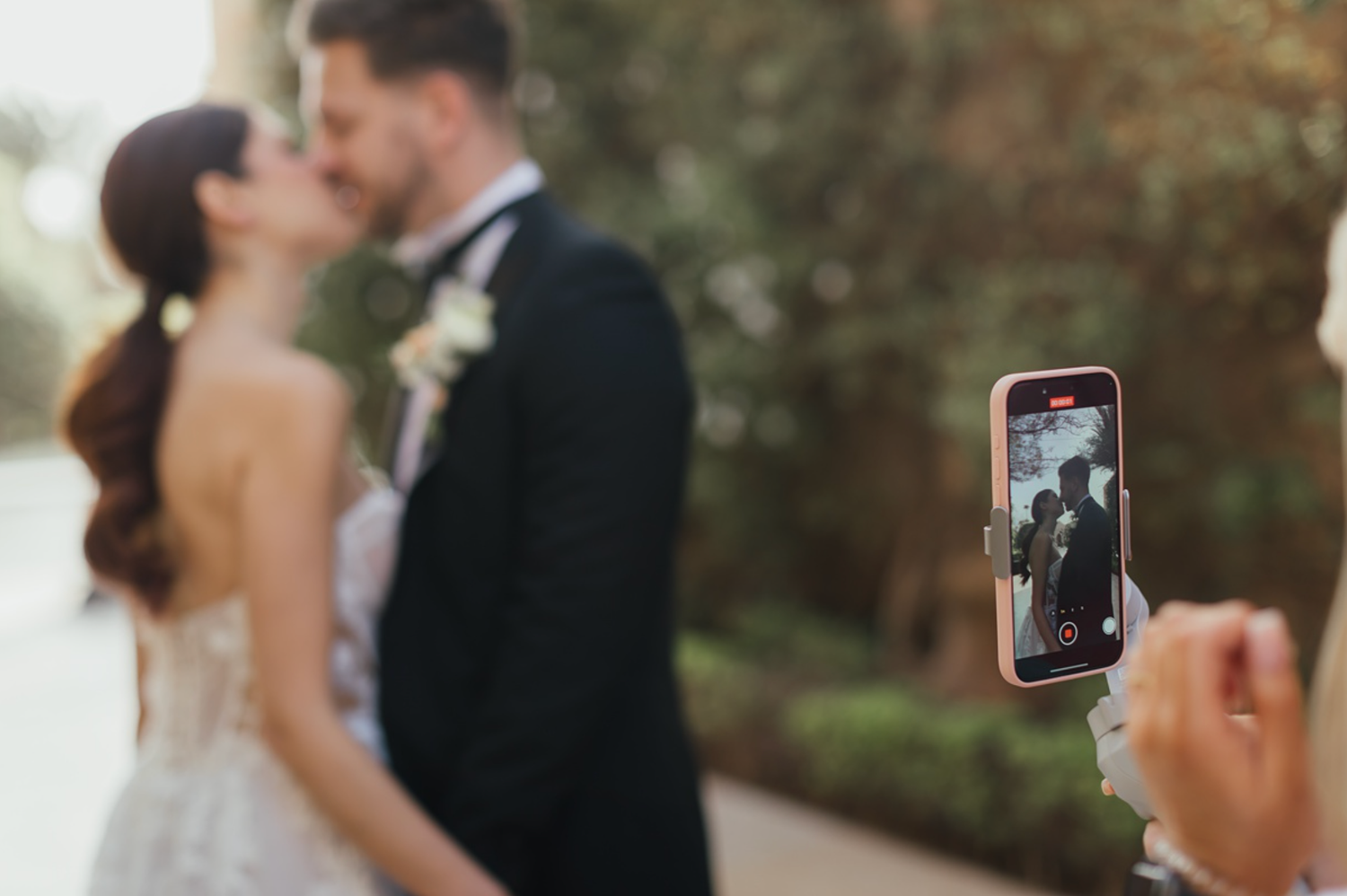 A phone captures a couple kissing at their wedding, with the bride in white and groom in a tuxedo blurred in background.