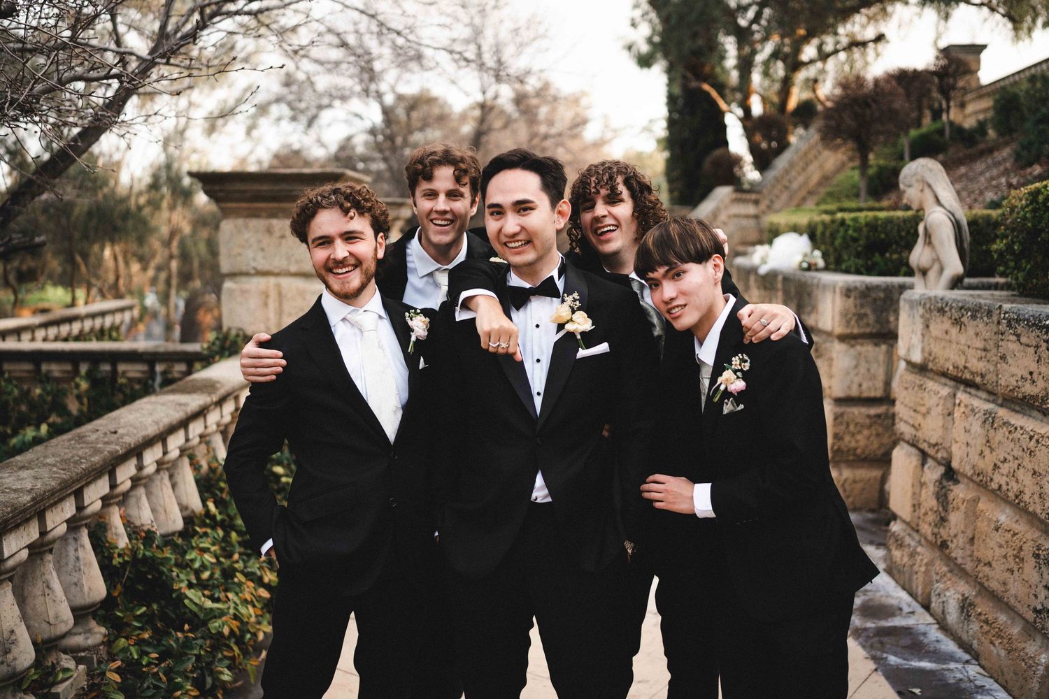 A group wearing black formal attire poses together outdoors by stone architecture at sunset.