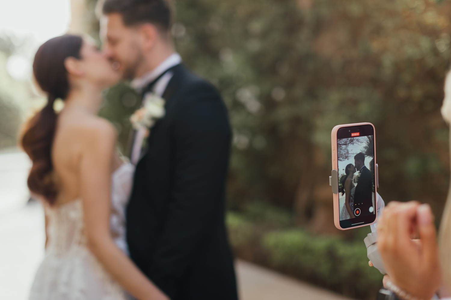 A smartphone captures a couple kissing at their wedding, with the bride in white and groom in a tuxedo blurred in the background.