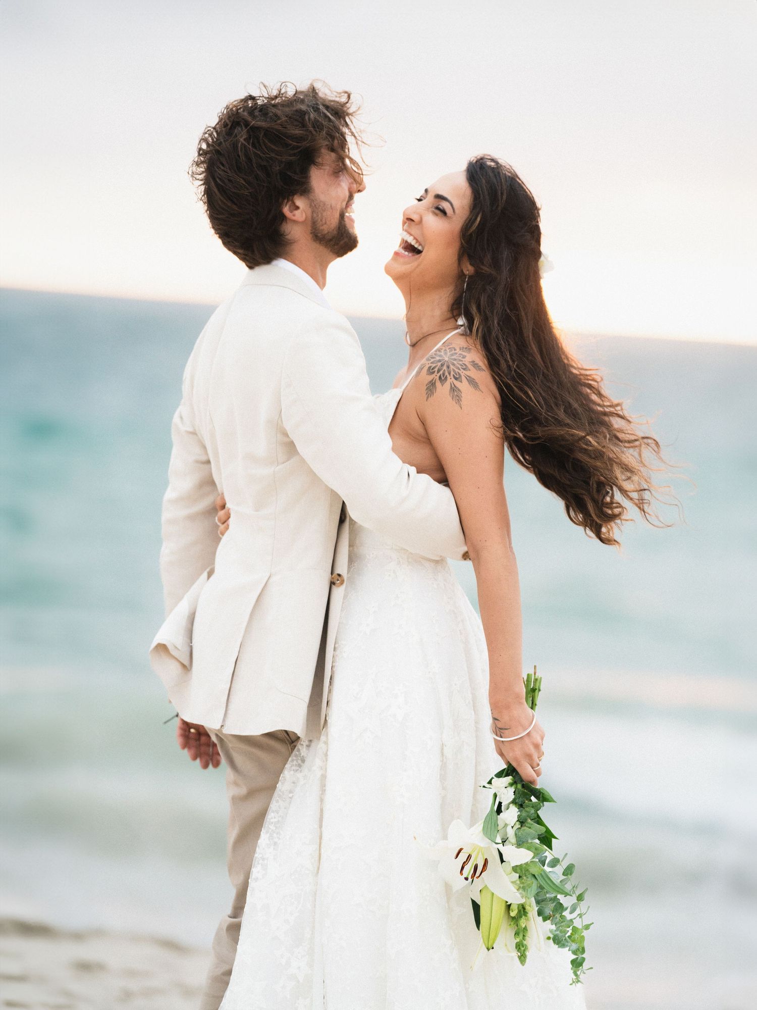Candid beach wedding photos of couple in white outfits celebrating by the ocean.