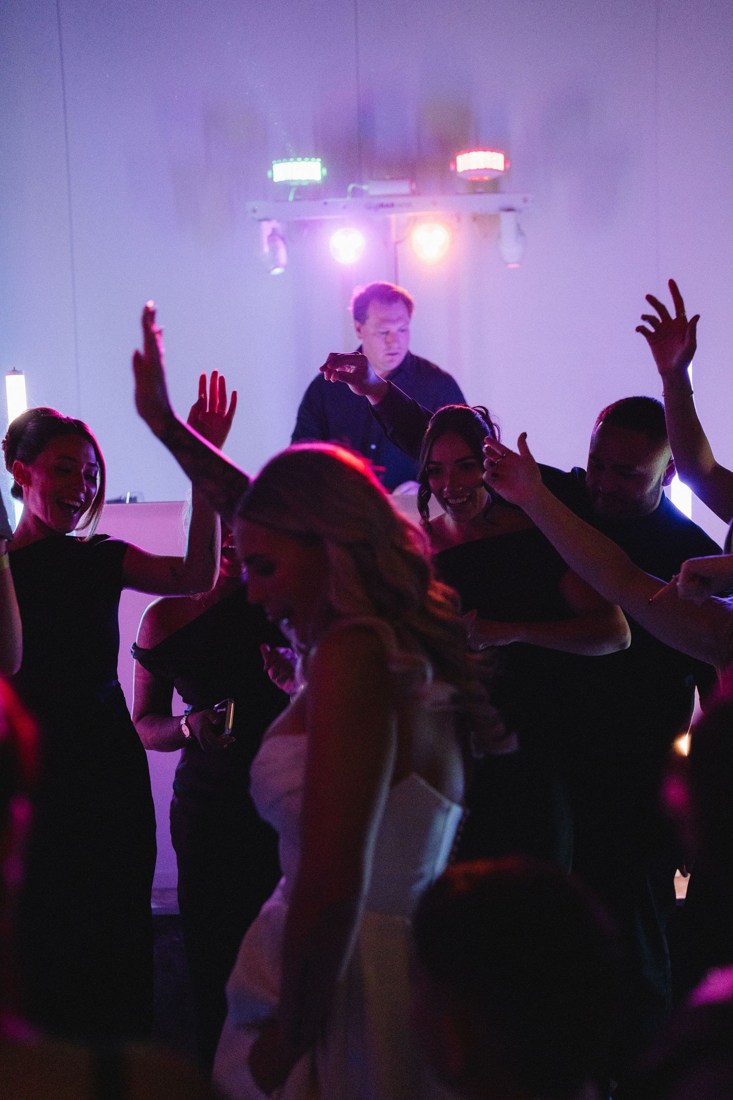 Wedding guests dancing with hands raised at a lively reception party, with a DJ performing on stage under colorful lights.