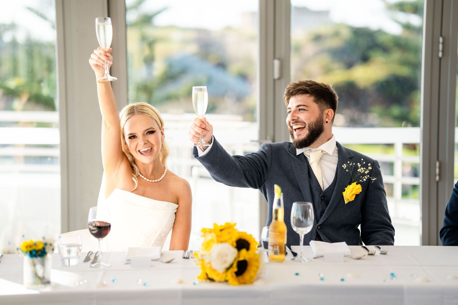 Wedding reception scene with celebratory toasts and sunflowers decorating a bright indoor venue.
