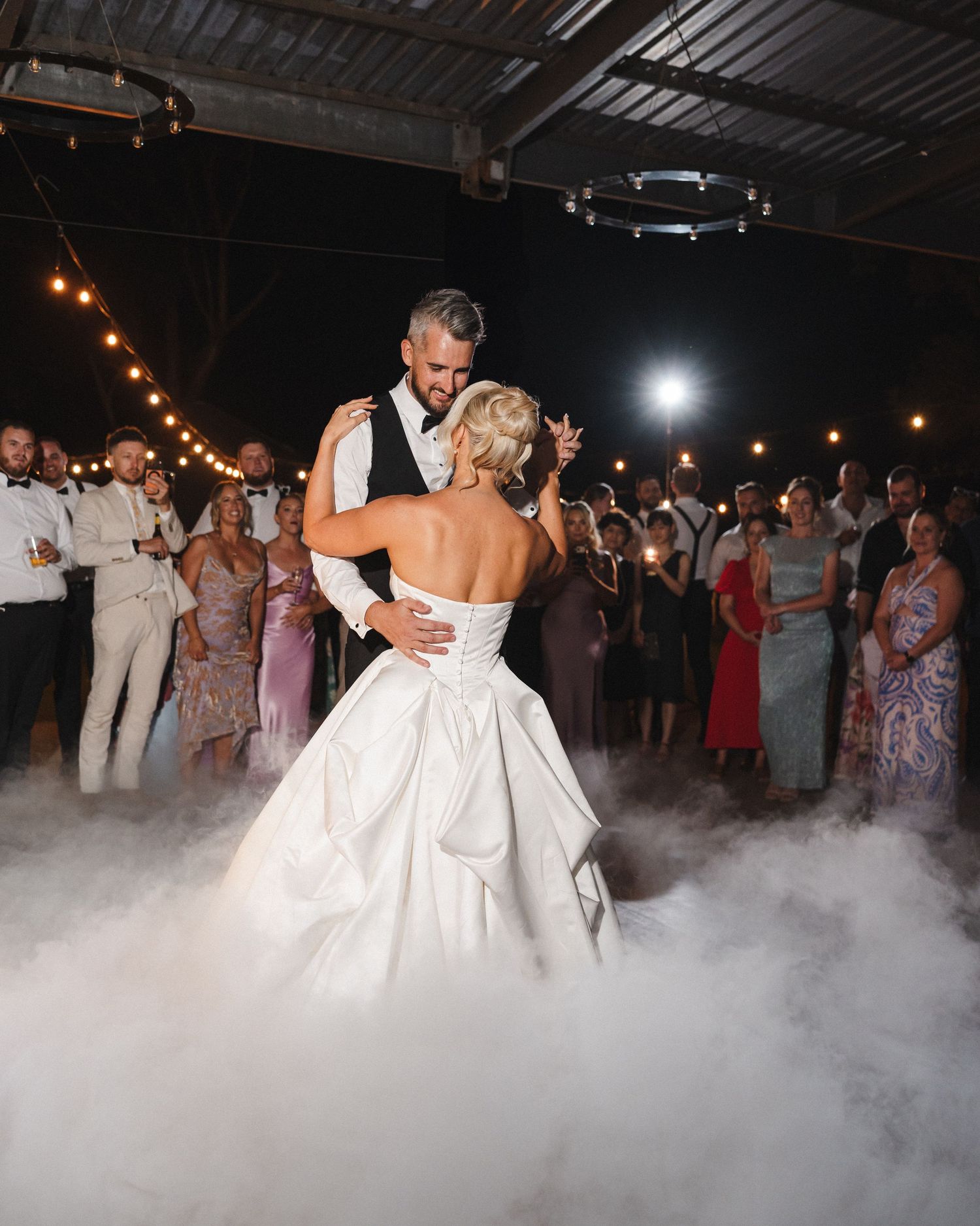 Bride and groom share first dance surrounded by guests and string lights on a foggy dance floor.