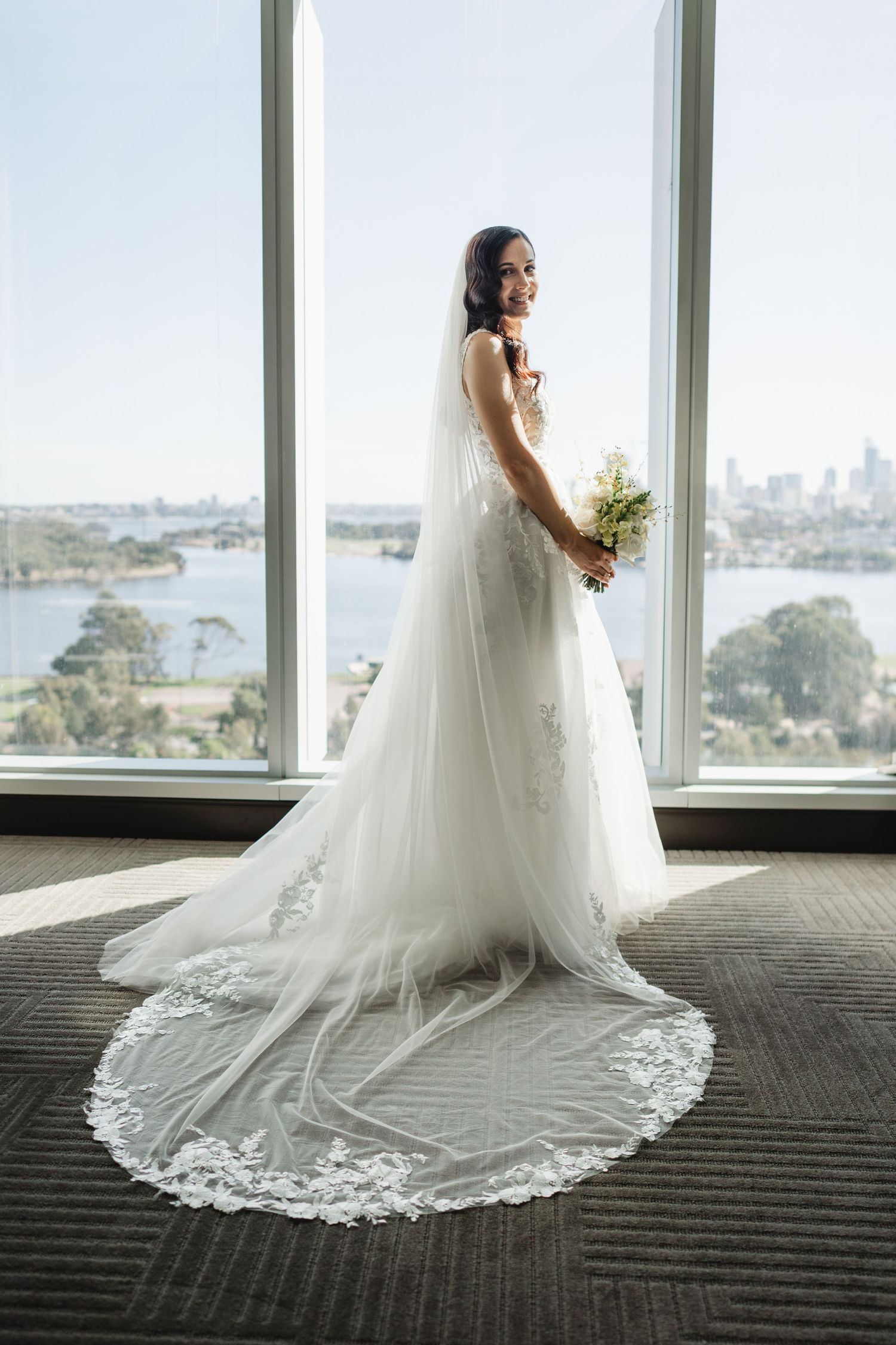Bride in elegant white lace wedding gown with long train and veil, holding bouquet, standing by large windows with city view.