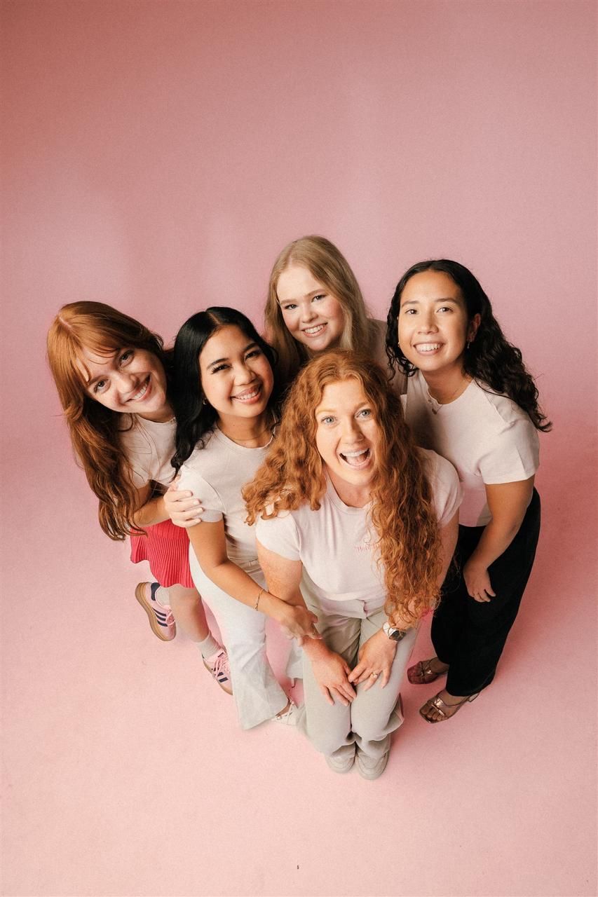 Five diverse women in white tops smiling together against a pink background, photographed from above.
