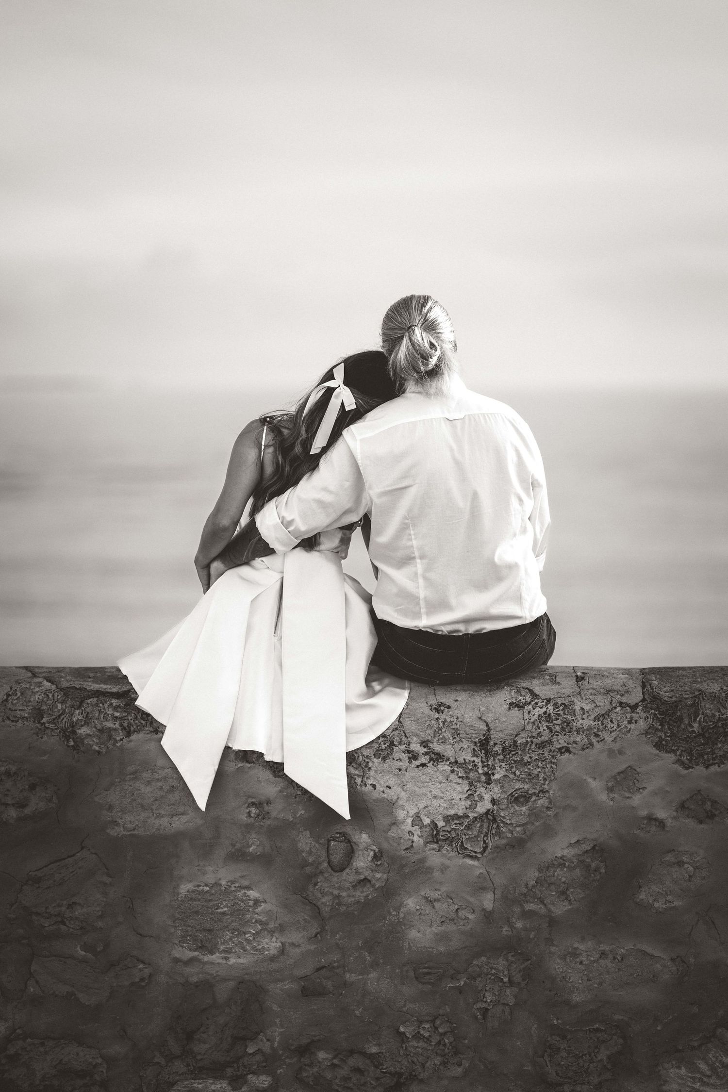 Artistic black and white portrait of wedding couple sitting on rocky terrain against cloudy sky.