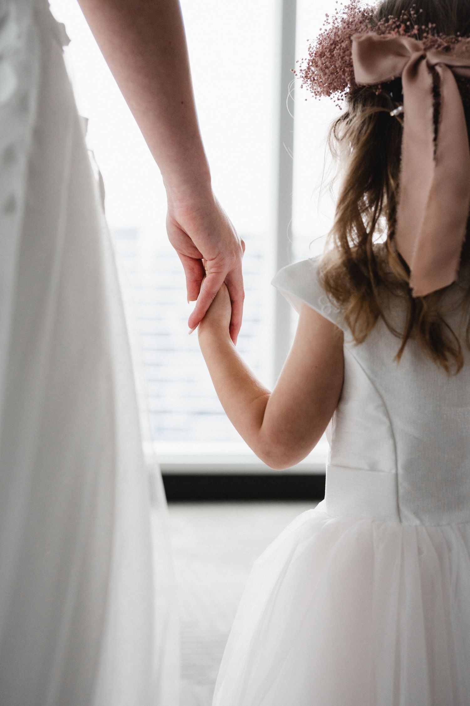A young flower girl in a white dress and floral bow holds an adult's hand near a bridal gown by a window.