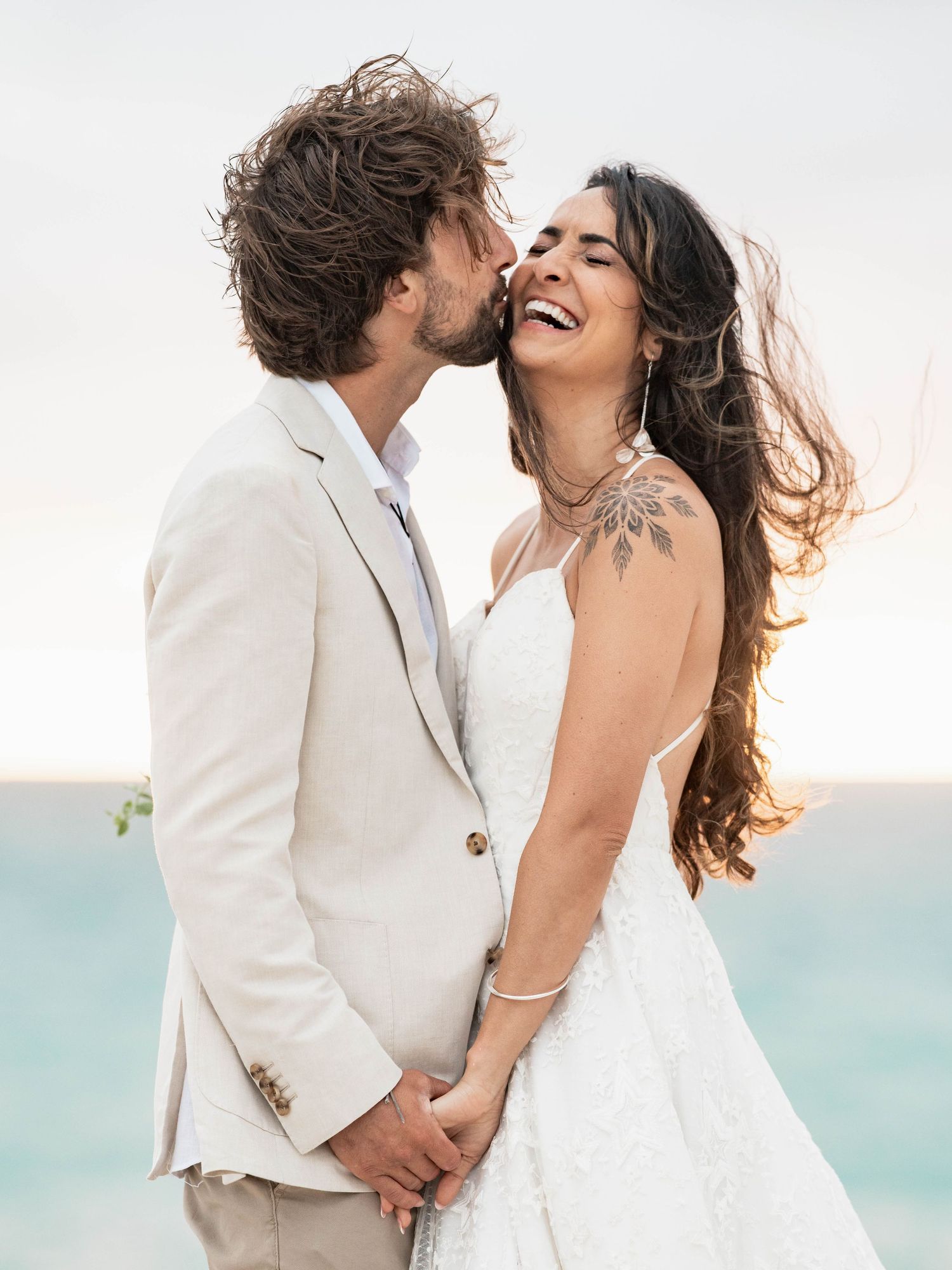 Romantic beachside wedding photos show couple embracing in white attire against turquoise water.