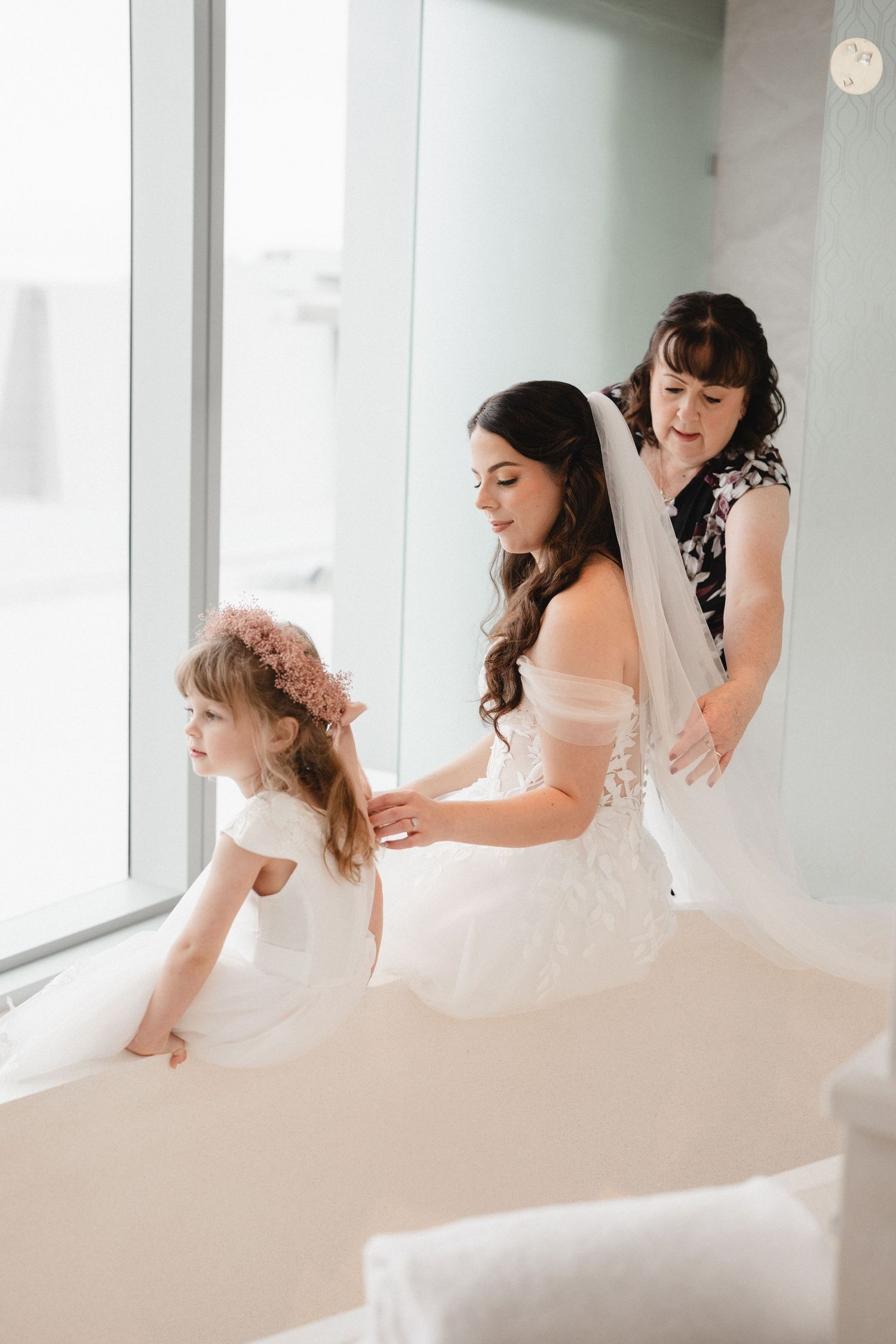 Bride in white gown gets veil adjusted by woman while flower girl in white dress sits nearby by large window.