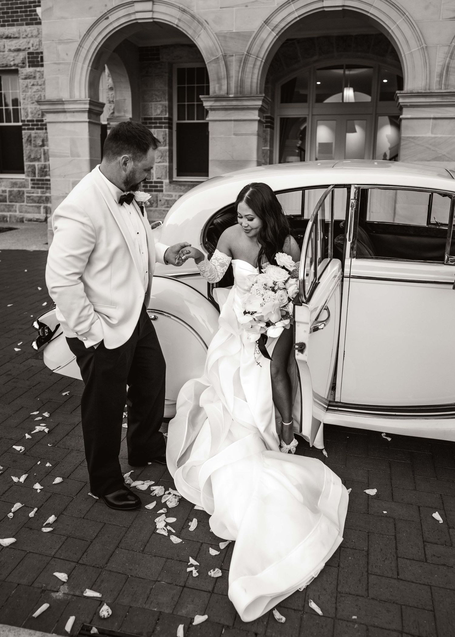 Wedding couple entering vintage car with white dress and rose petals on ground.