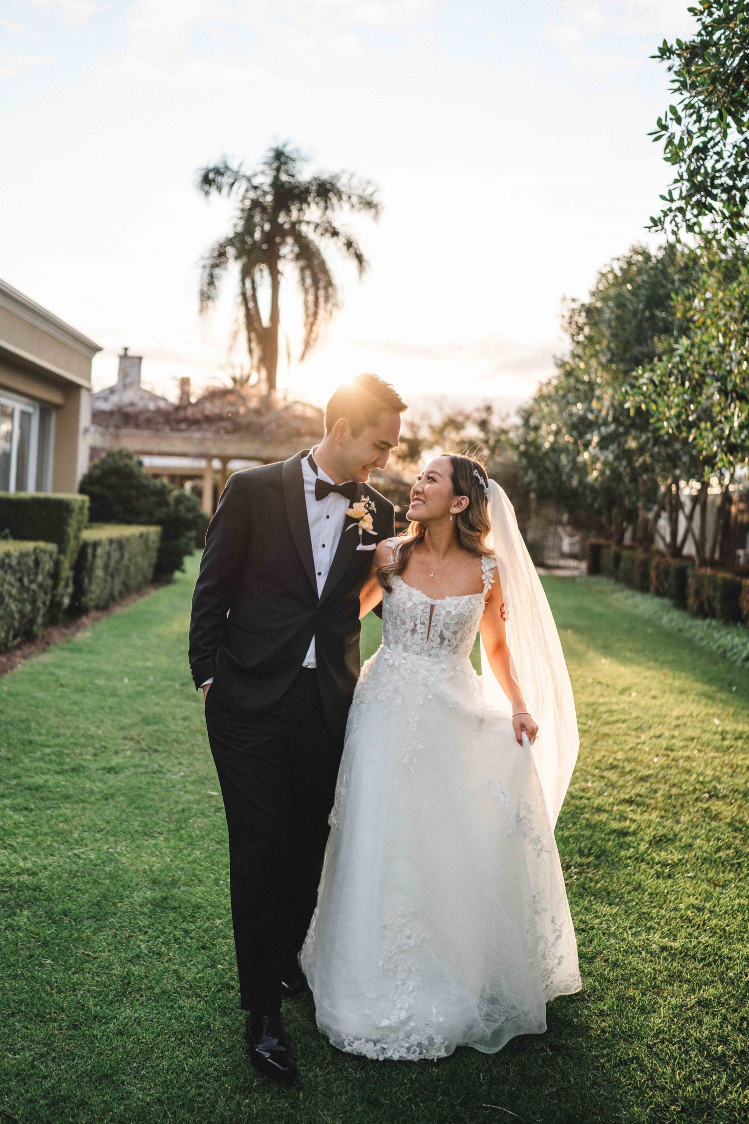 Happy couple in wedding attire share intimate moments on a sunlit lawn with palm trees in the background.