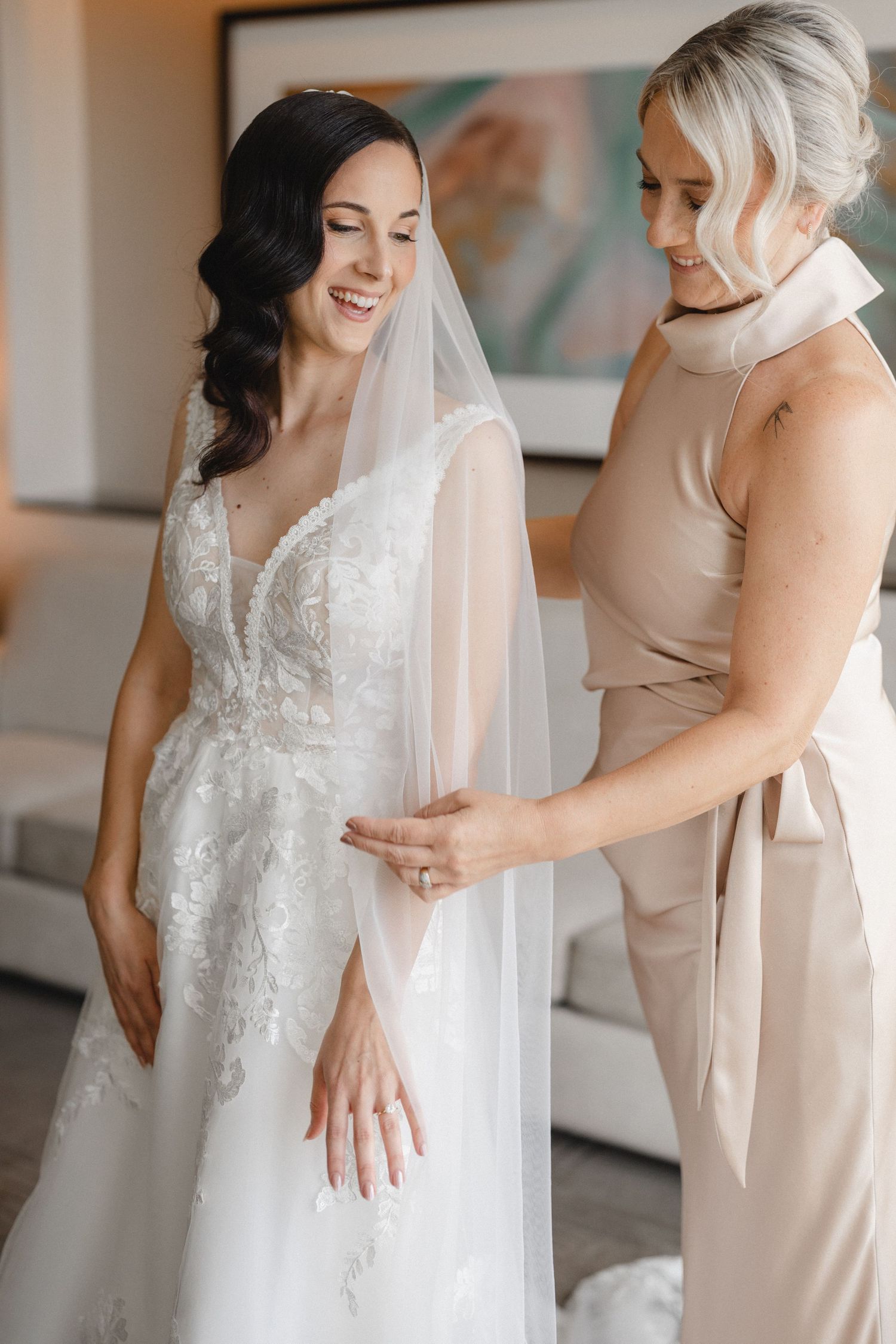 Bride in lace wedding gown and veil smiles as bridesmaid in champagne dress helps her get ready.