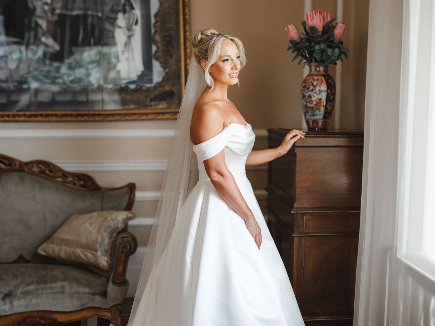 Bride in off-shoulder white ball gown with veil poses elegantly in a classic interior room.