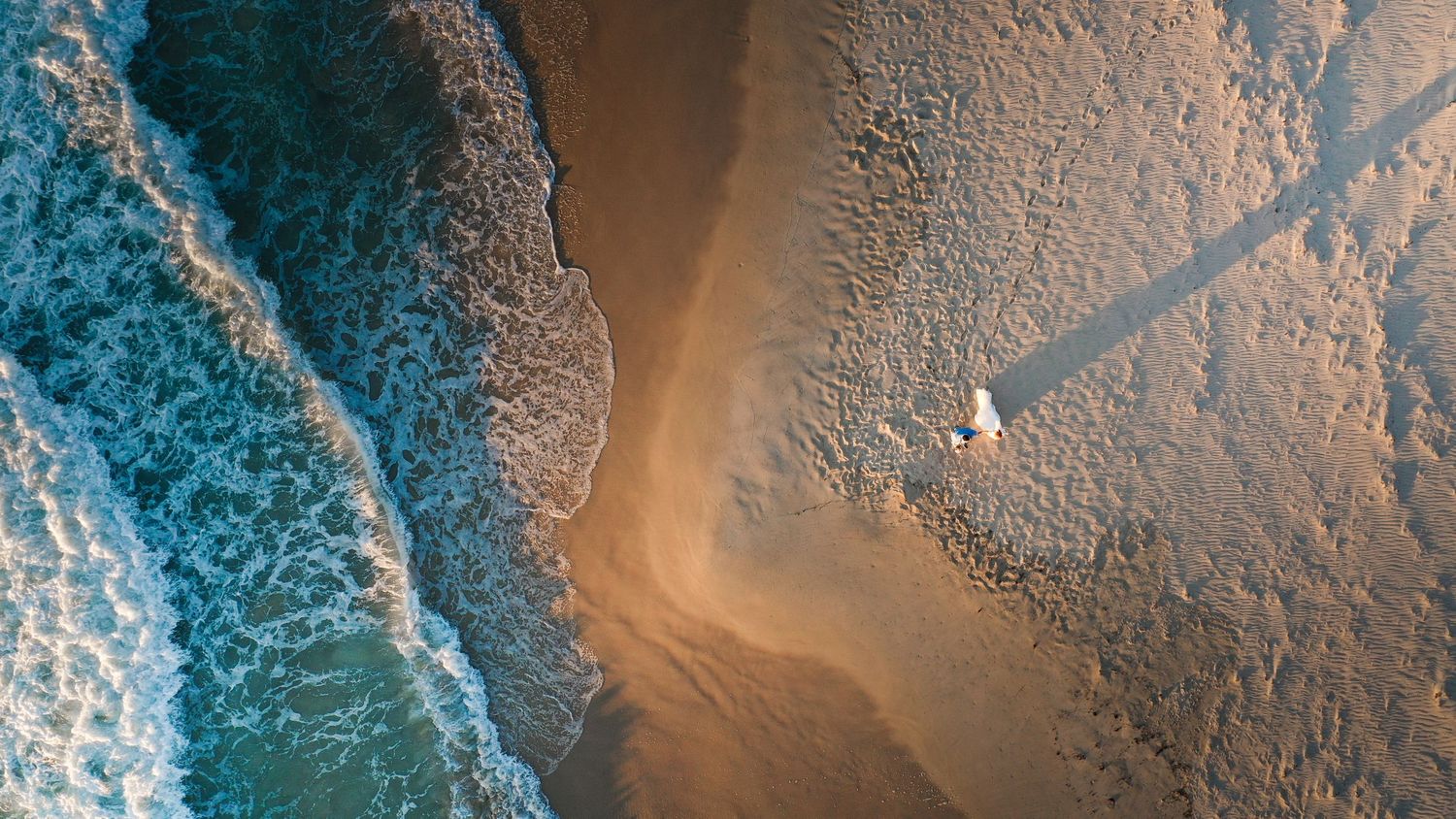 Aerial view of turquoise ocean waves crashing on a sandy beach with a lone figure walking along the shoreline.