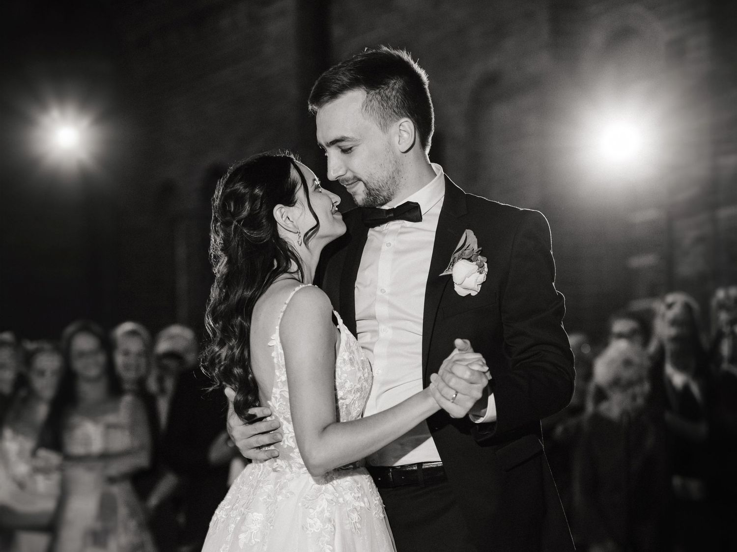 Black and white wedding photo of bride and groom sharing first dance, surrounded by guests in elegant venue.