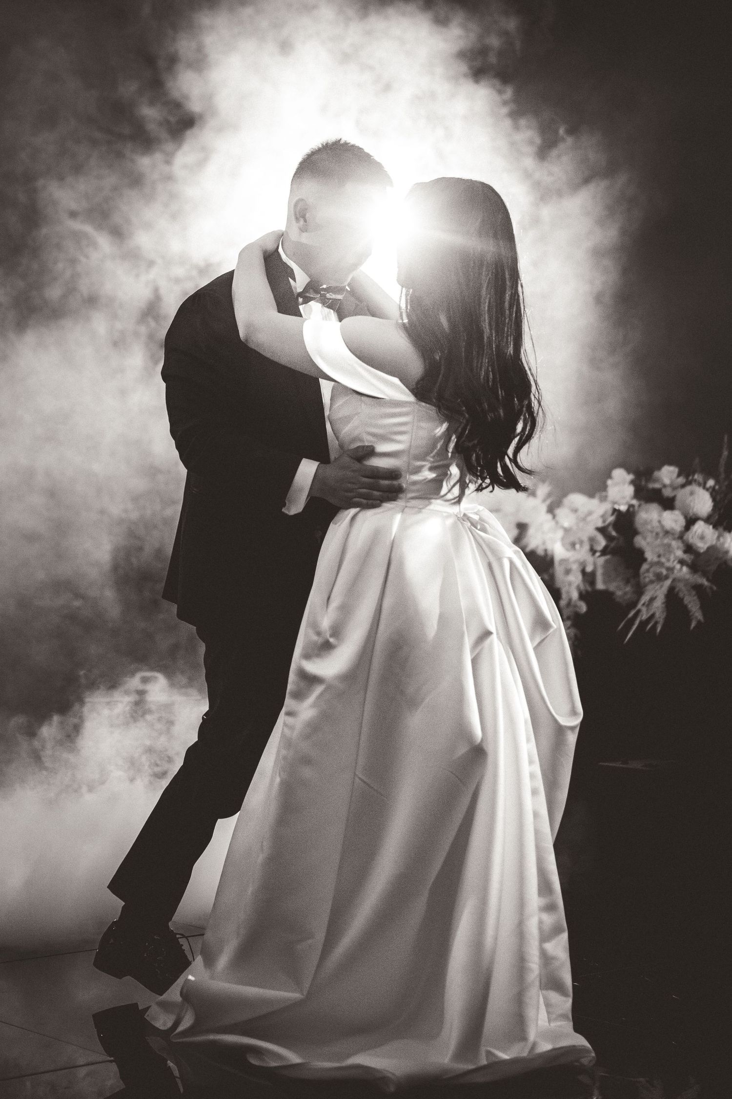 Romantic black and white wedding photo with dramatic backlighting and fog creating a dreamy silhouette of newlyweds.