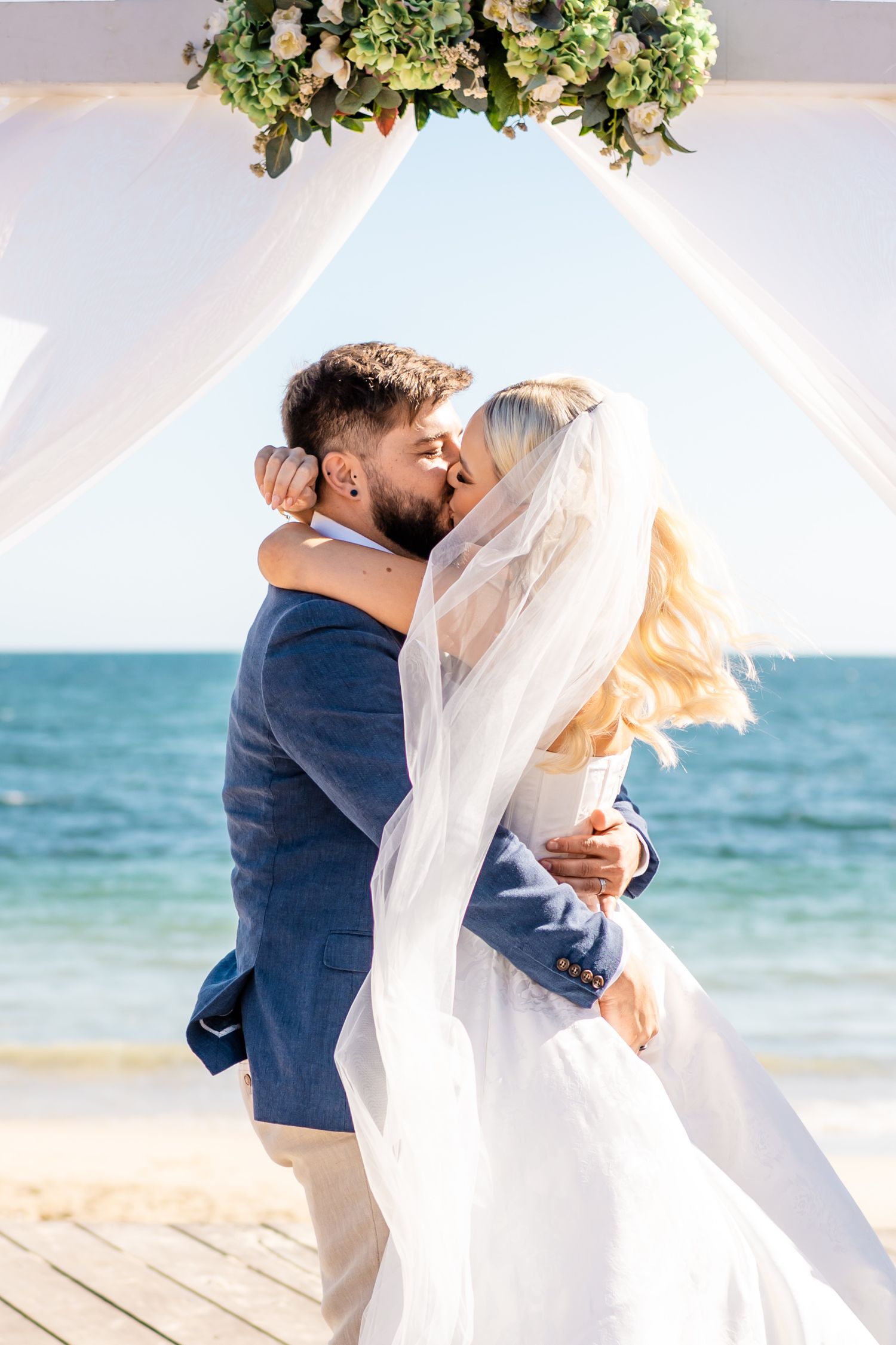 Romantic beach wedding kiss under a floral arch with ocean backdrop and flowing veil in the breeze.