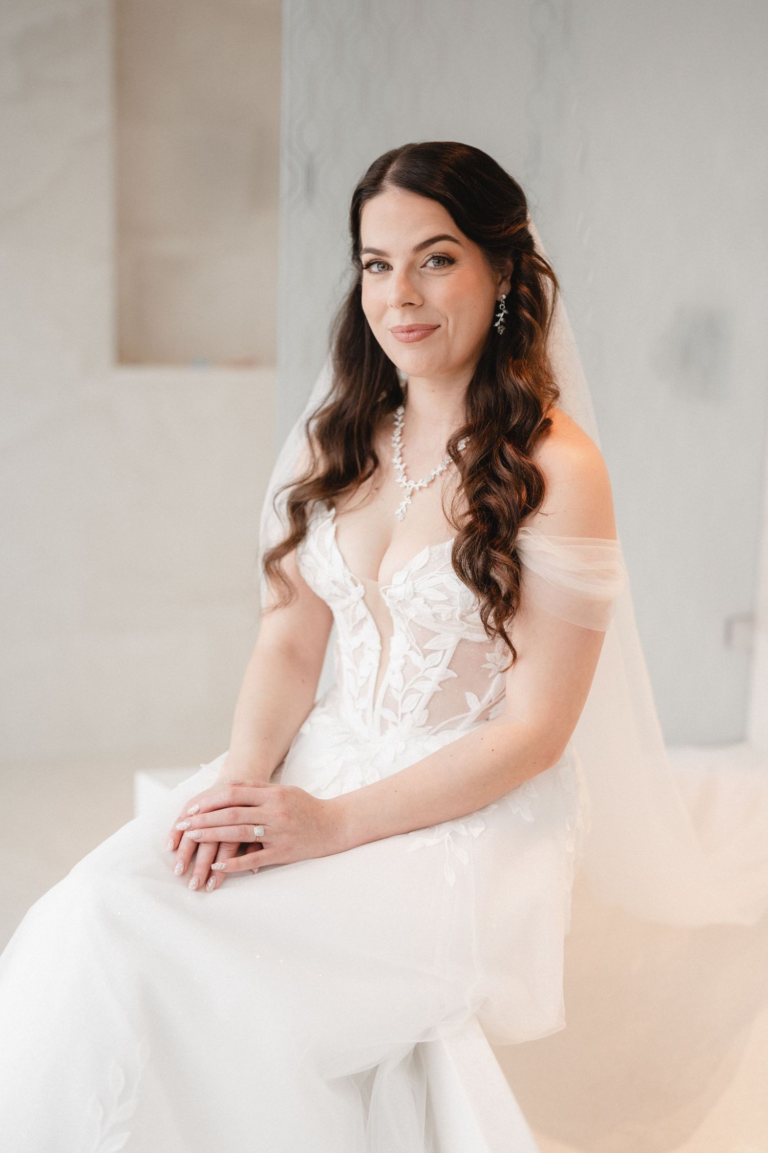Elegant bride in off-shoulder lace wedding dress with veil, pearl necklace, and wavy brunette hair, seated in bright room.