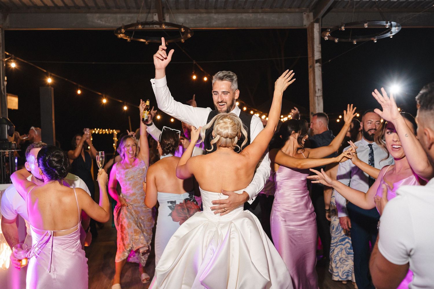 Wedding guests dance together on a crowded dance floor under colorful purple lighting at an evening reception celebration.