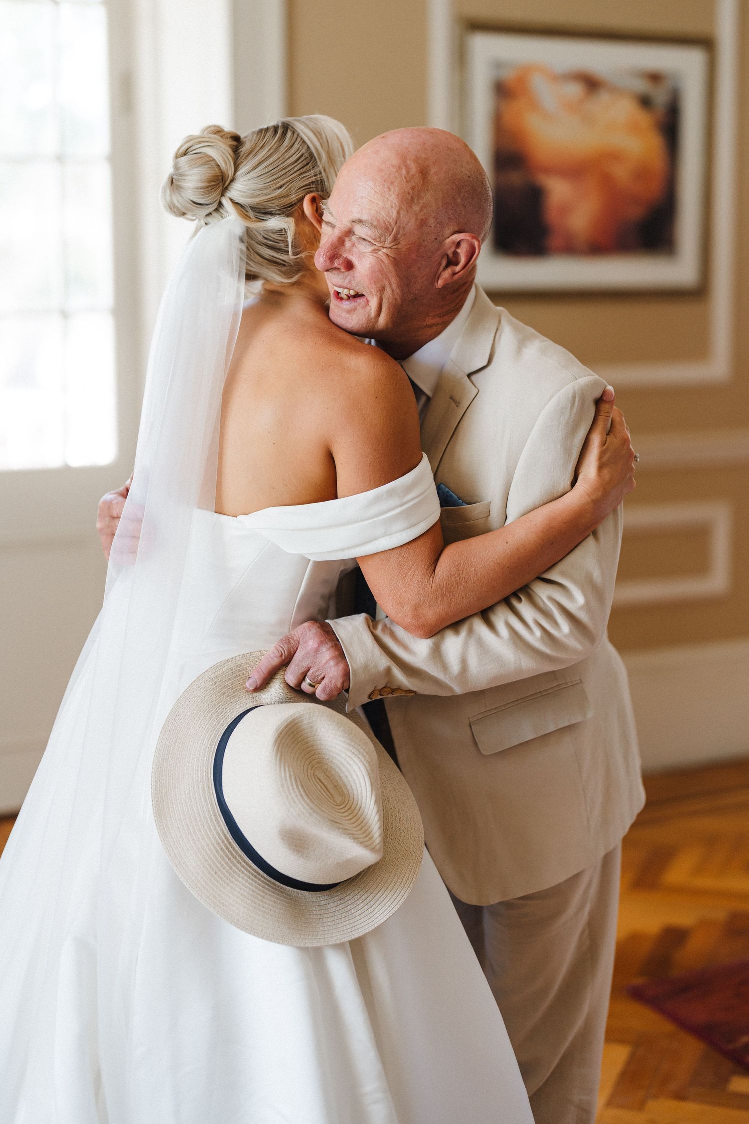 A tender moment captured as a bride in a white gown embraces someone wearing a light beige suit and hat.