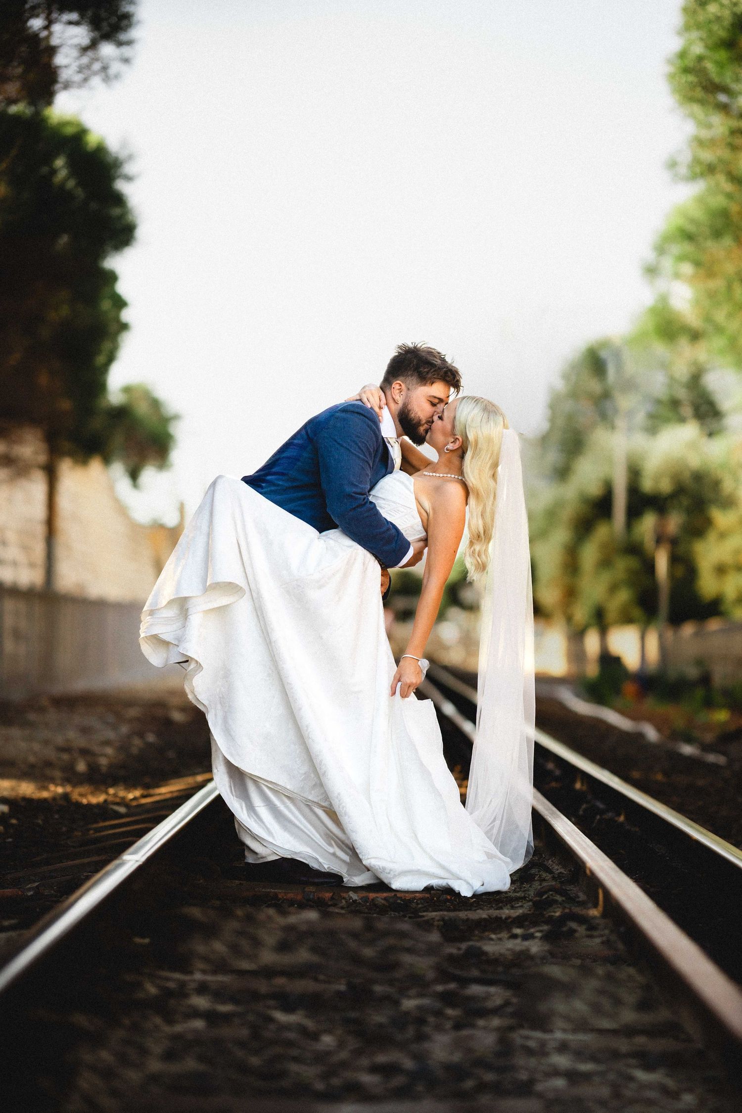 Dramatic wedding photo of couple sharing romantic dip kiss on railroad tracks.