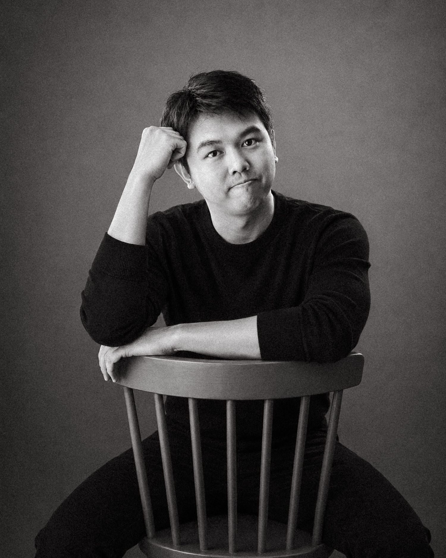 Black and white portrait of a young Asian man in a black sweater, seated on a wooden chair, resting his chin thoughtfully.