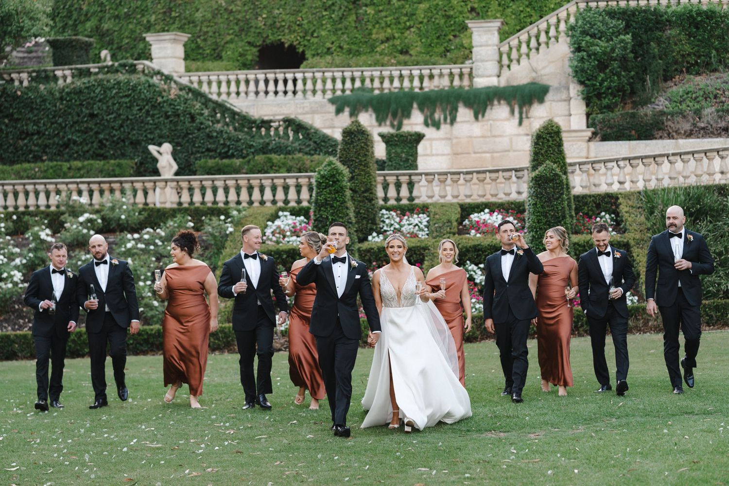 Wedding party in formal attire poses on manicured lawn with elegant stone balustrade and greenery in background.