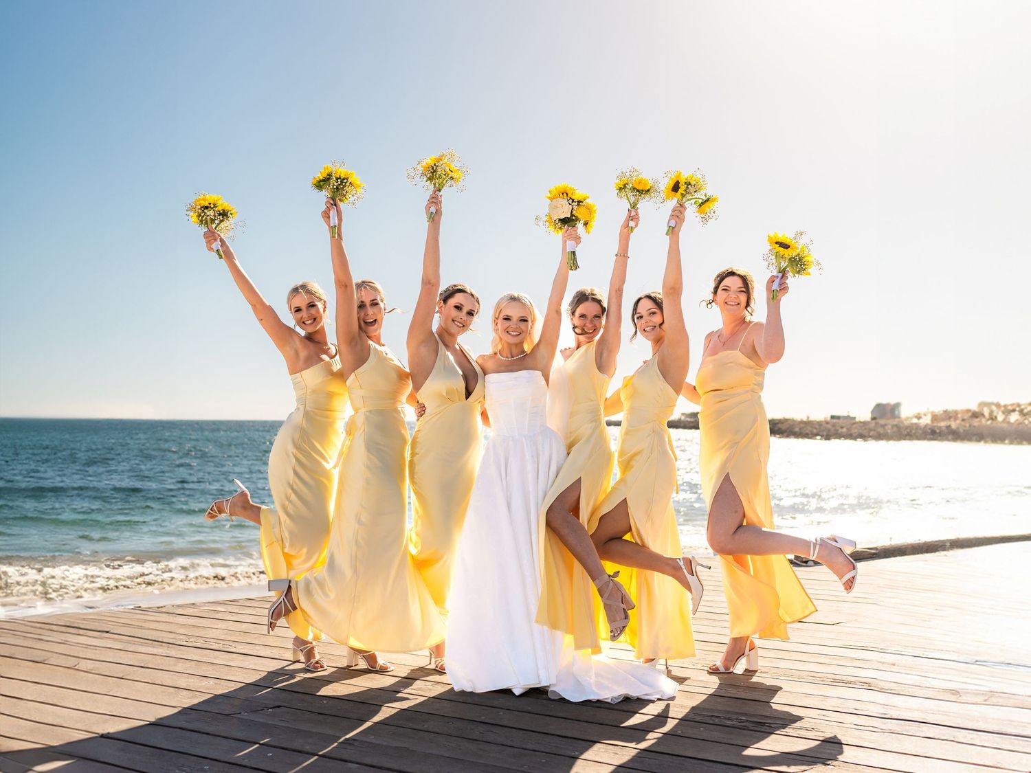 Bride in white gown jumps with six bridesmaids in yellow dresses holding sunflower bouquets on a sunny beachside pier.