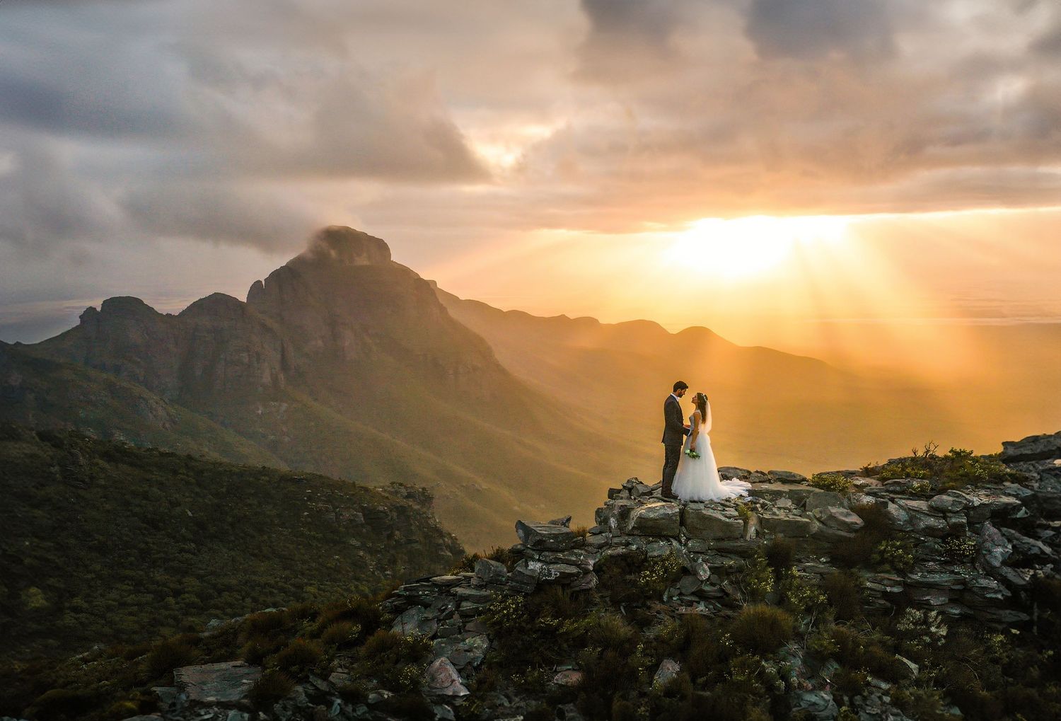 Bride and groom stand on rocky mountain peak at golden sunset with dramatic rays of light and green valleys below.