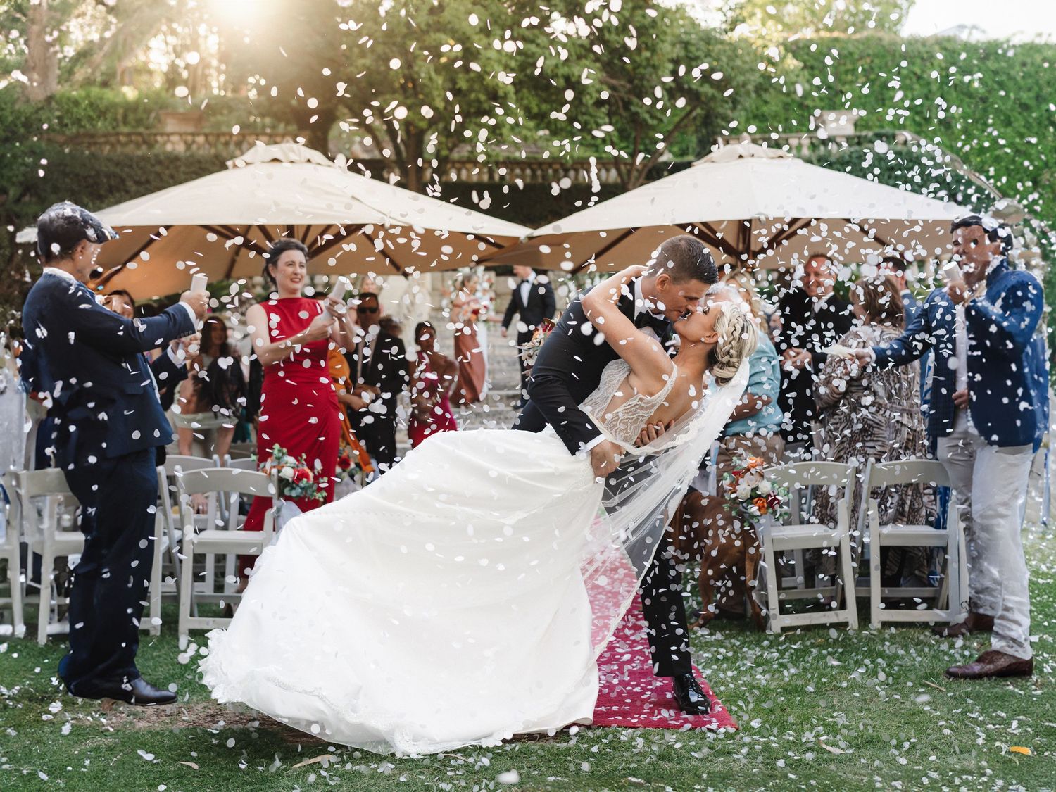 Newlywed couple sharing first dance kiss surrounded by confetti at outdoor wedding reception.
