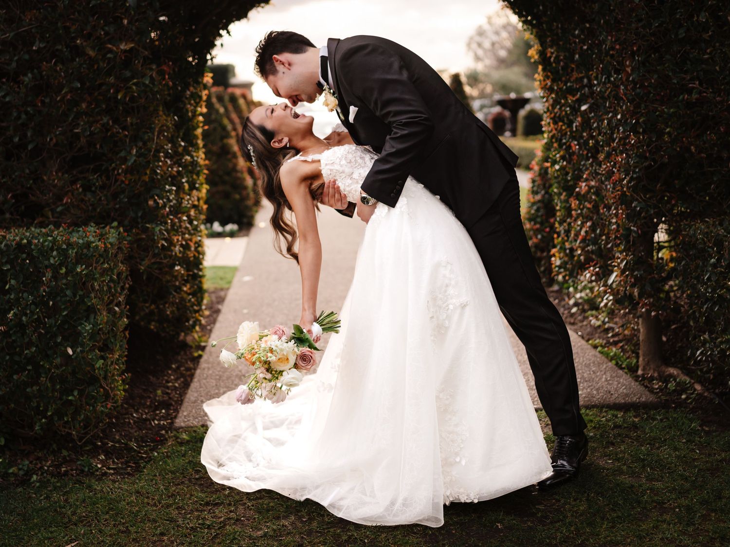 Romantic garden wedding photo with couple sharing dramatic dip kiss under archway.